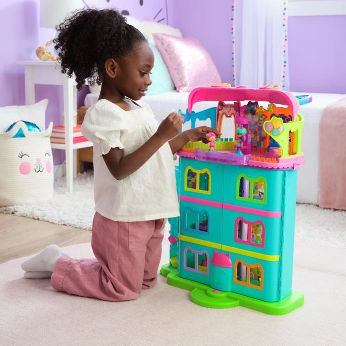 Una bambina gioca con una casa delle bambole colorata, inginocchiata su un tappeto. La casa ha più livelli e finestre. Camera da letto e giocattoli visibili.