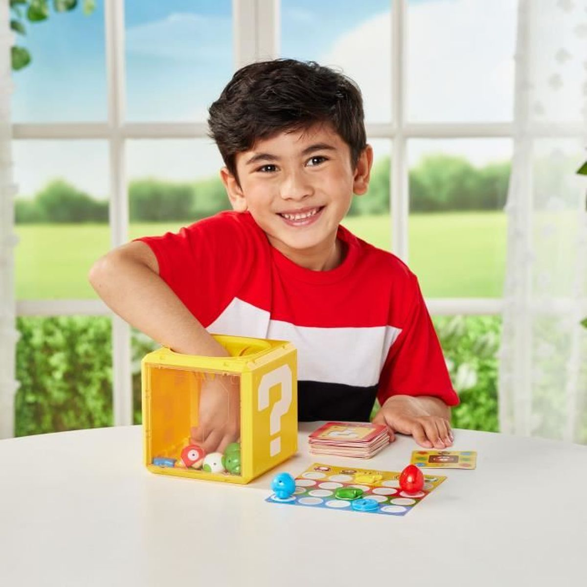 Een lachende jongen in een rood shirt grijpt in een gele doos met een vraagteken. Spelstukken en kaarten op tafel.