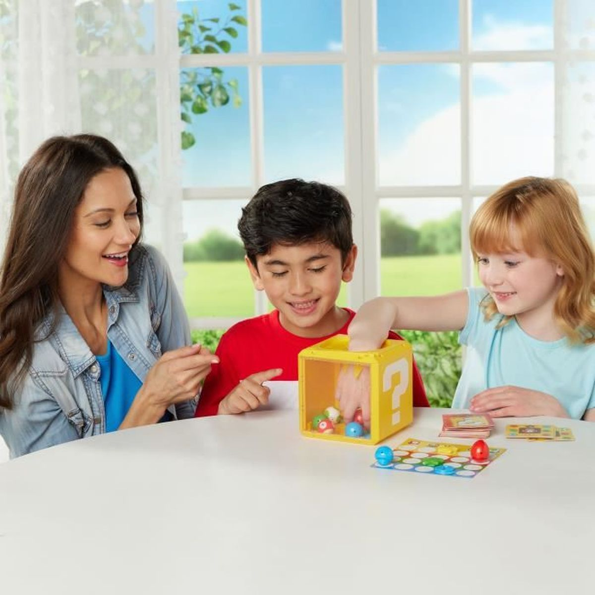 Een vrouw en twee kinderen spelen een spel met een gele doos. Gekleurde ballen, kaarten en een bord liggen op een witte tafel.