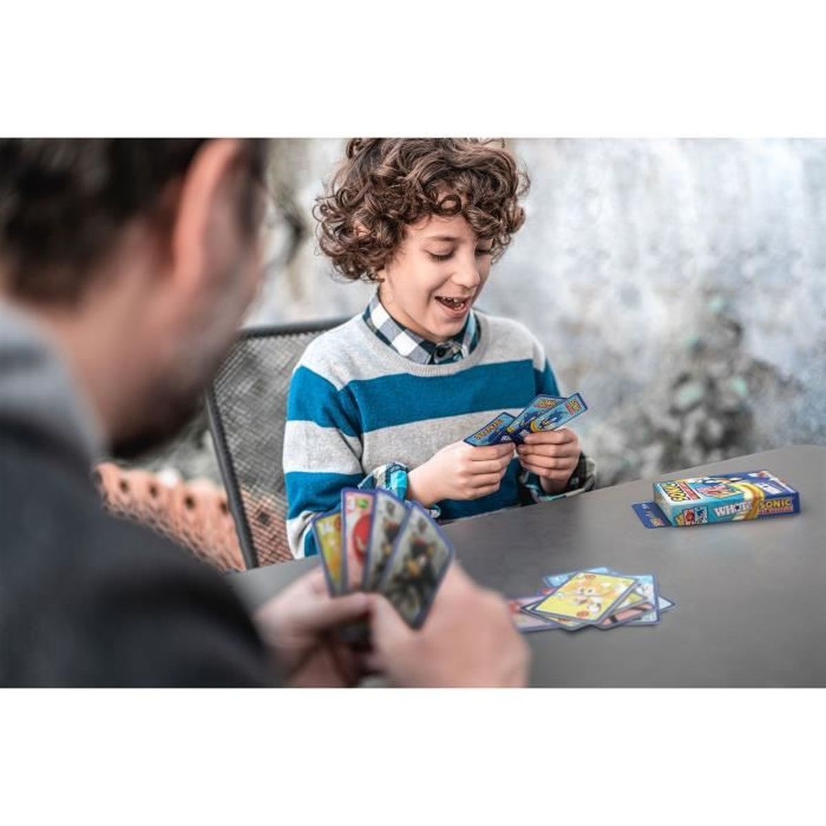 Chłopiec i osoba grają w karty. Chłopiec uśmiecha się i trzyma karty. Pudełko do gry karcianej leży na stole.