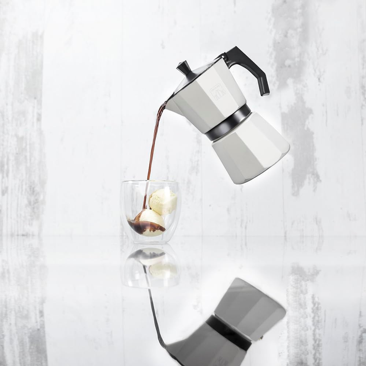 Eine weiße Espressomaschine gießt Kaffee in ein Glas mit Eis. Die Szene spiegelt sich. Weißer Holz-Hintergrund.