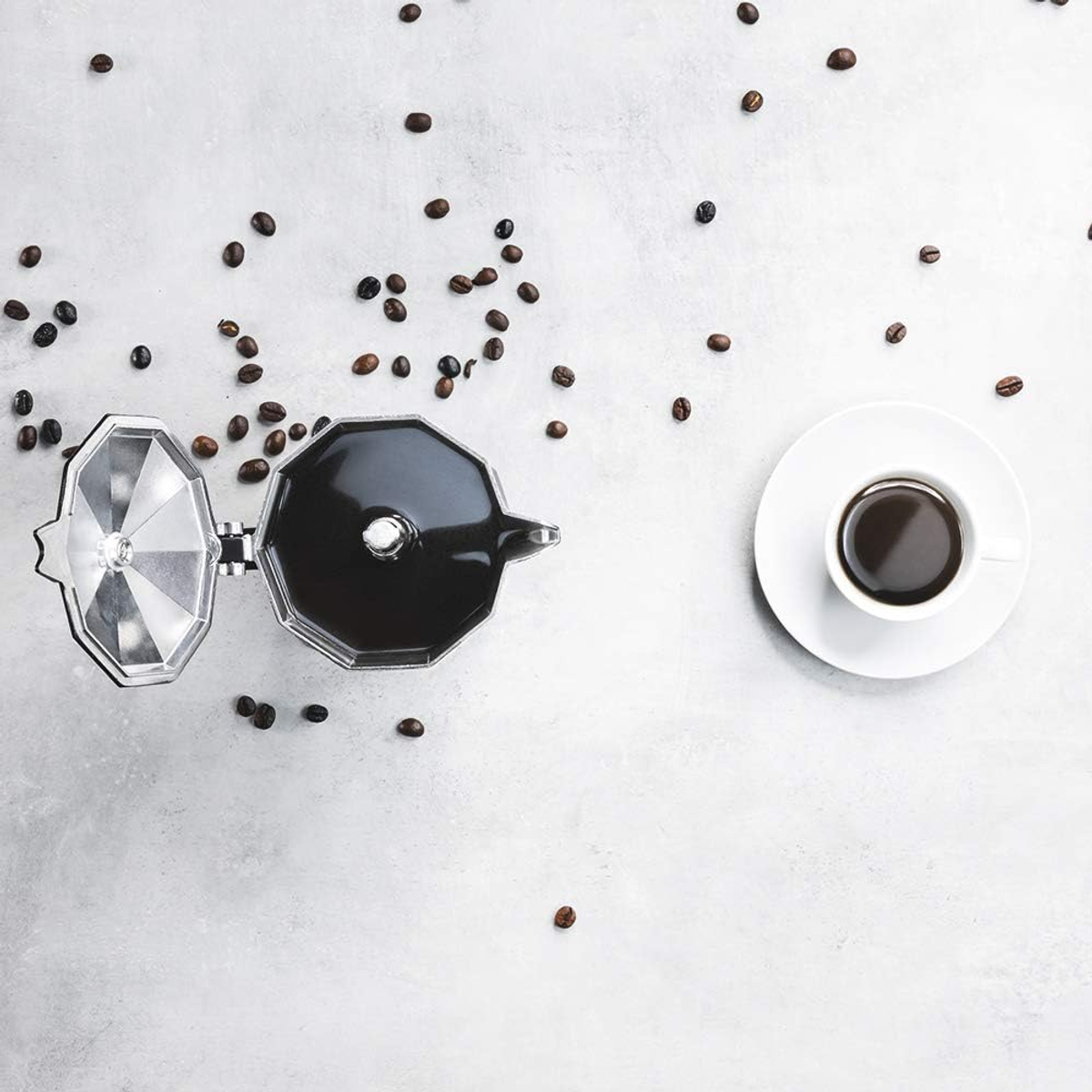Draufsicht auf Kaffeebohnen, die um eine schwarze Kaffeemaschine und eine Tasse Kaffee auf einer weißen Untertasse verstreut sind.