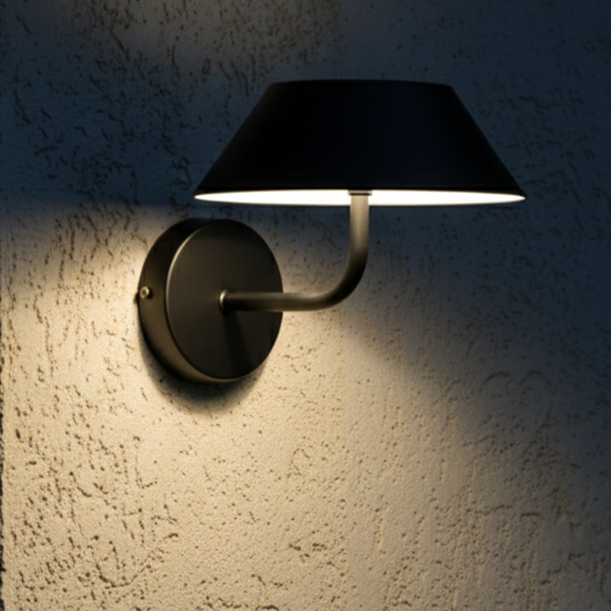 Eine schwarze Wandleuchte mit Lampenschirm an einer strukturierten Wand. Die Lampe strahlt warmes Licht aus.