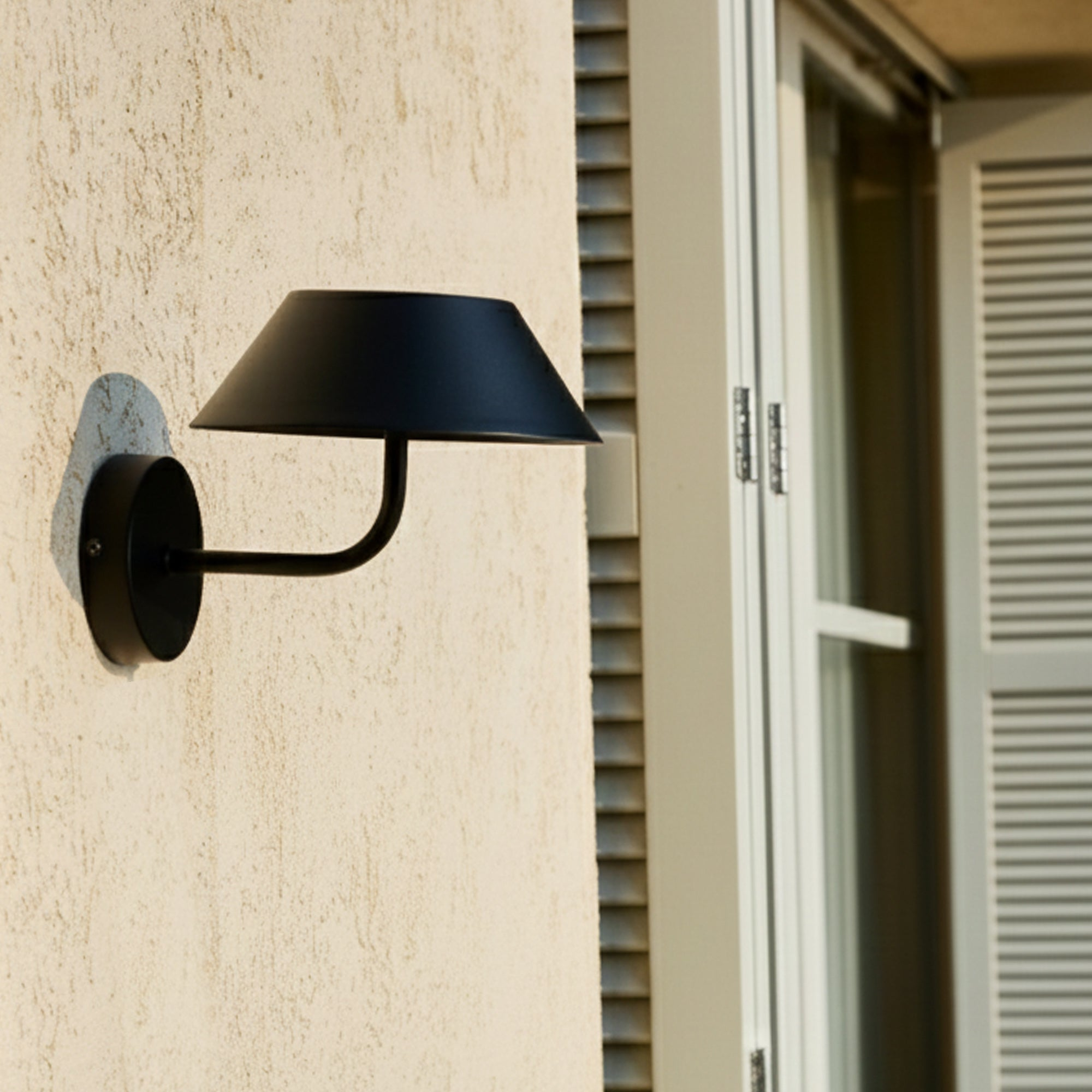 Eine schwarze Außenlampe ist an einer strukturierten beigefarbenen Wand befestigt. Rechts befindet sich ein Fenster mit weißen Fensterläden.