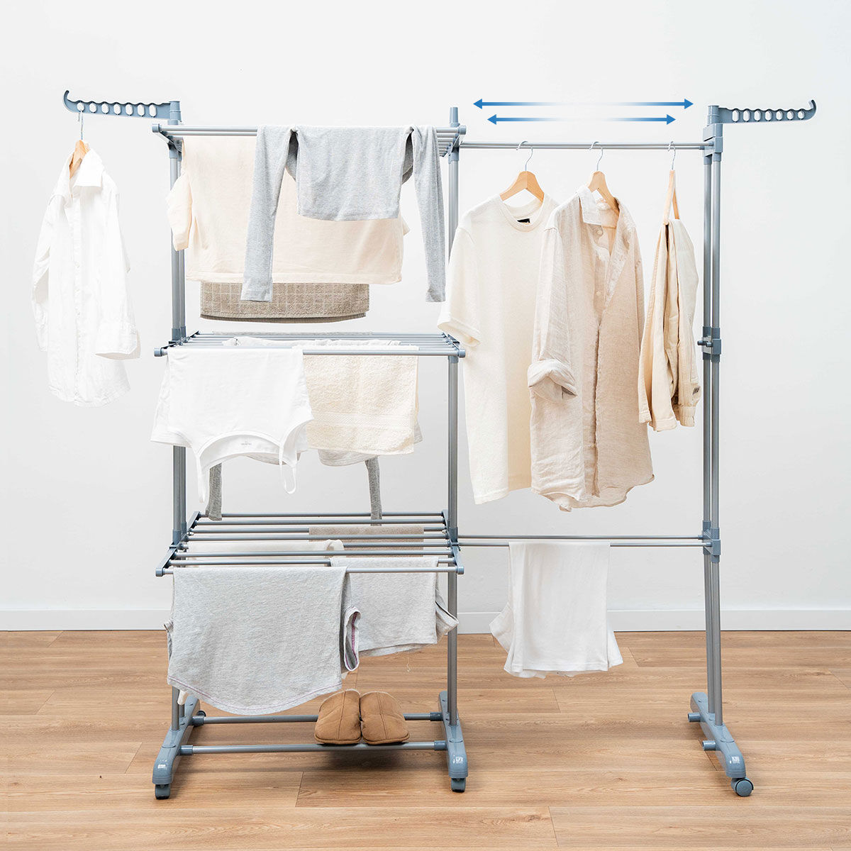 Ein Wäscheständer mit verschiedenen Kleidungsstücken. Enthält Hemden, Handtücher und Hosen, vor weißer Wand und Holzboden.