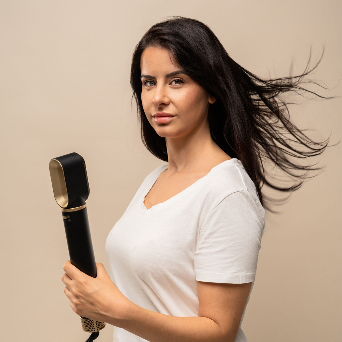 Eine Frau hält ein schwarzes Haarstyling-Werkzeug. Ihr dunkles Haar weht, sie trägt ein weißes Hemd und schaut in die Kamera. Beigefarbener Hintergrund.