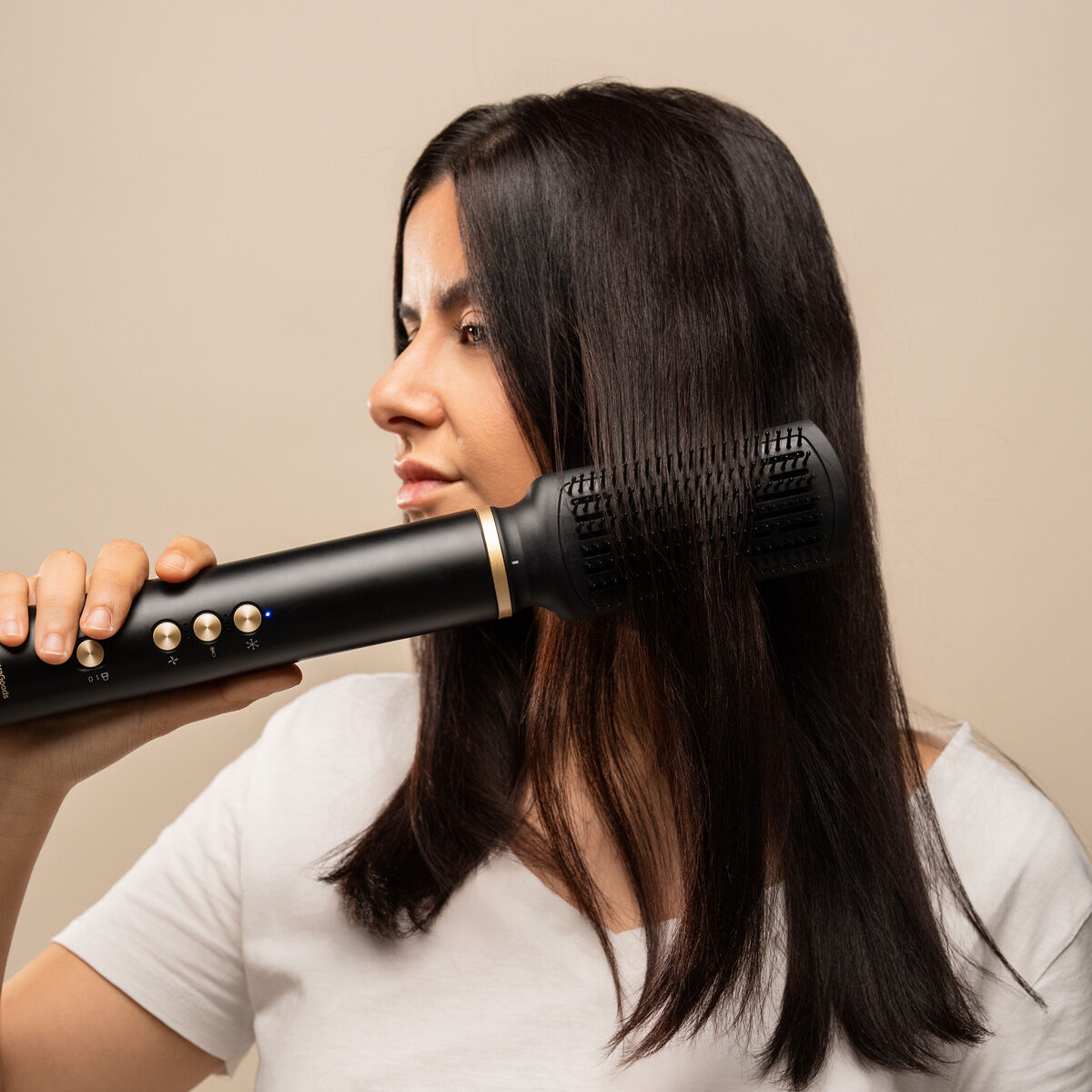 Eine Frau benutzt ein schwarzes Haarstyling-Werkzeug. Sie hat lange, dunkle Haare und trägt ein weißes Hemd vor beigem Hintergrund.