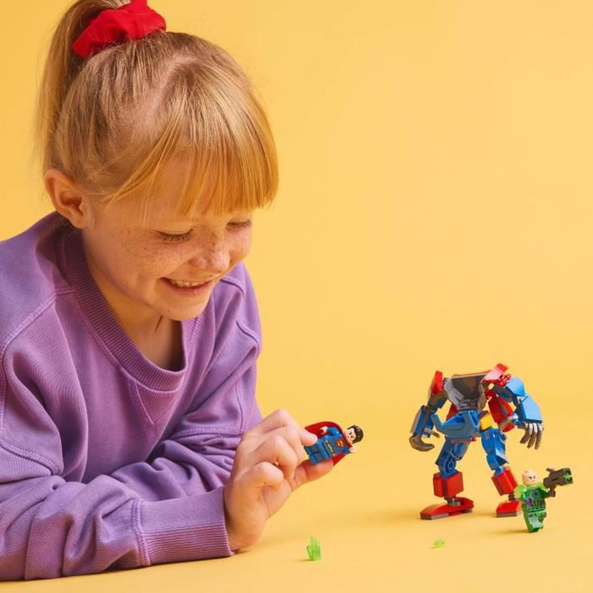 Dziewczyna uśmiecha się, bawiąc się superbohaterami Lego. Figurka Supermana w dłoni. Robot Lego i inne figurki na żółtej powierzchni.