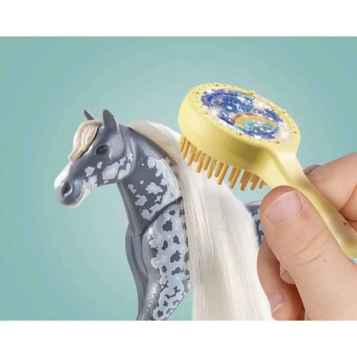 Ein Spielzeugpferd wird mit einer gelben Bürste gebürstet. Das Pferd ist grau mit weißen Flecken und hat eine weiße Mähne. Die Bürste hat ein farbenfrohes Design.