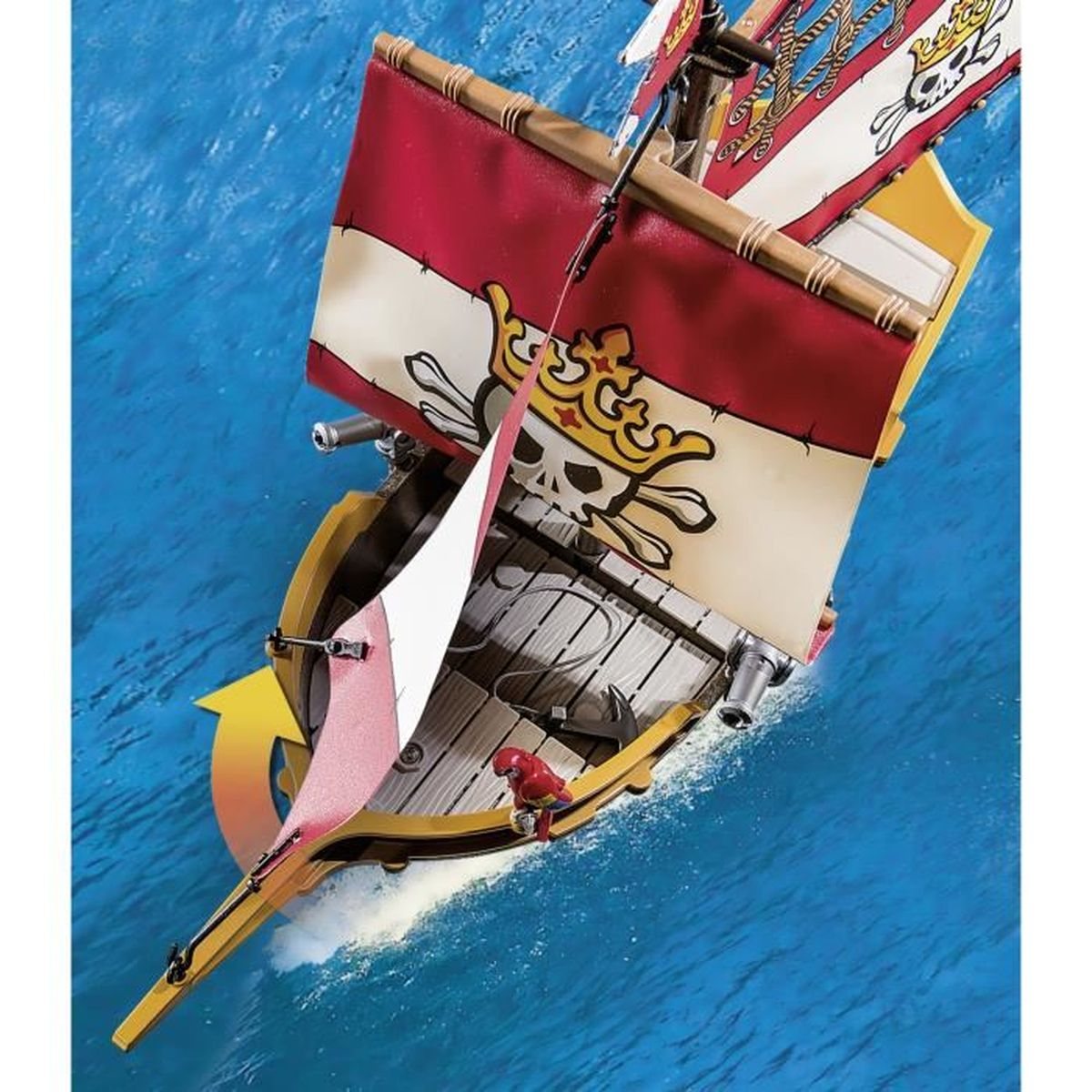 Una nave pirata giocattolo naviga sull'acqua blu. La nave ha vele rosse e bianche con un teschio e una corona. Un pappagallo rosso è a prua.