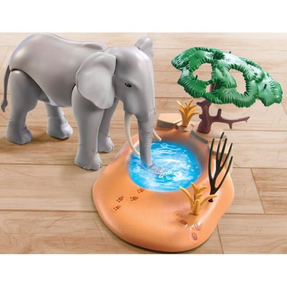 Ein grauer Spielzeug-Elefant trinkt aus einem blauen Wasserloch. Die Szene umfasst einen grünen Baum, gelbe Pflanzen und Fußabdrücke.