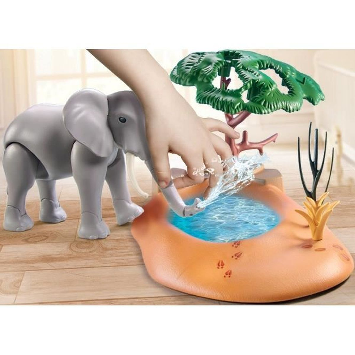 Ein Spielzeug-Elefant trinkt Wasser aus einem blauen Pool. Eine Hand sprüht Wasser. Ein Baum und eine Pflanze sind im Hintergrund.