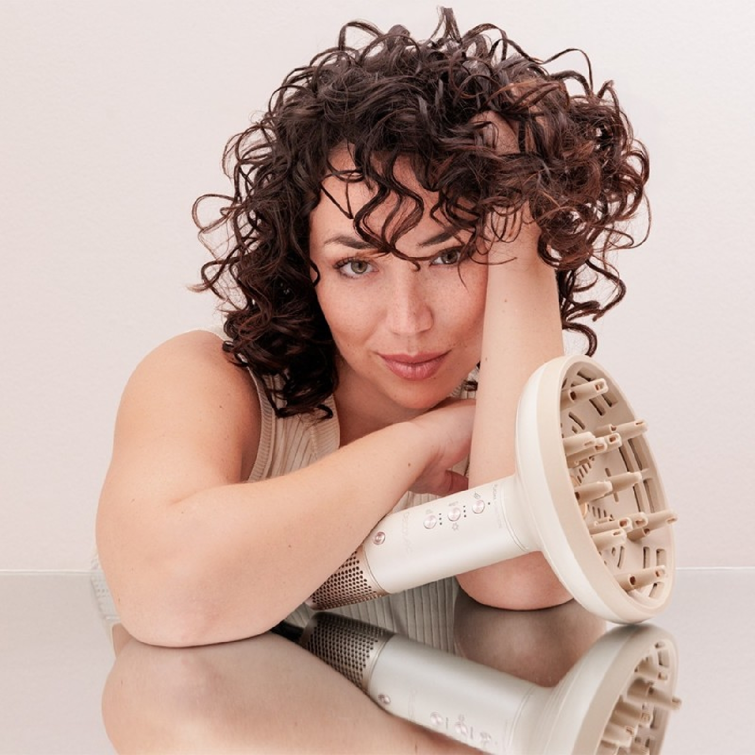 Kobieta z kręconymi włosami opiera się na stole. Trzyma suszarkę do włosów z dyfuzorem. Powierzchnia odbija obraz.