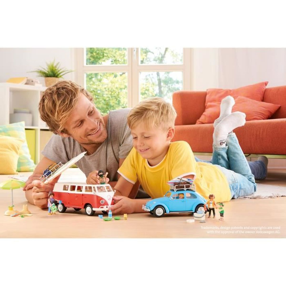Mężczyzna i chłopiec bawią się samochodzikami. Czerwono-biały van i niebieski samochód leżą na podłodze. W tle kanapa i okno.