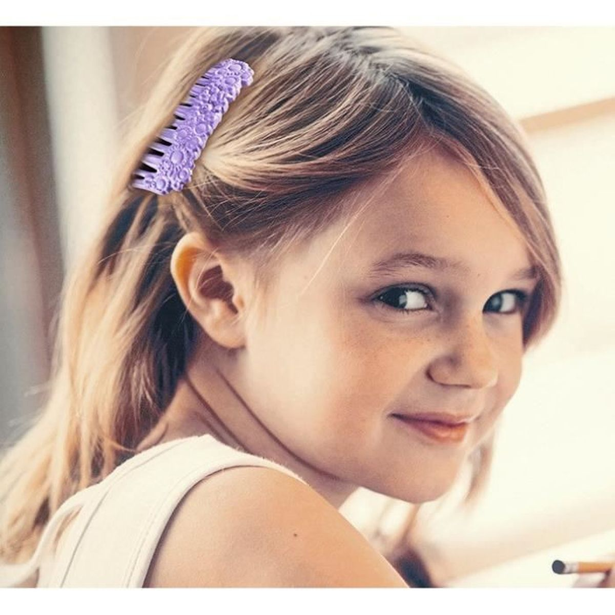 Una ragazza con i capelli biondi ha una forcina viola. Sta sorridendo. Indossa una maglietta bianca. Una matita è di lato.