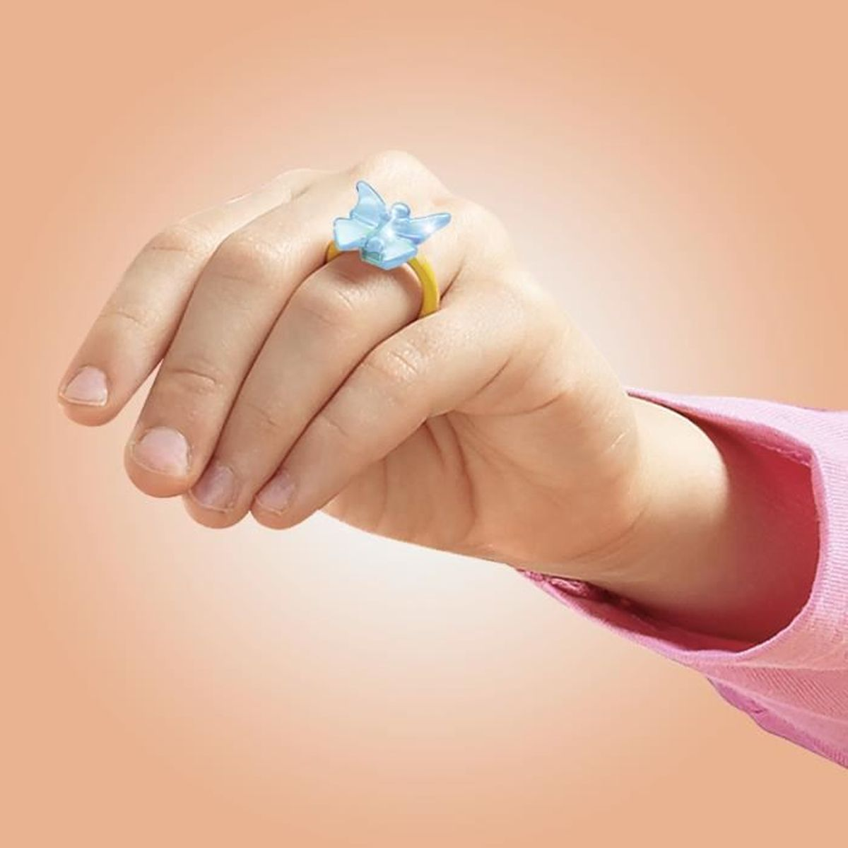 La mano di un bambino con un anello a farfalla blu con fascia gialla. Lo sfondo è color pesca chiaro.