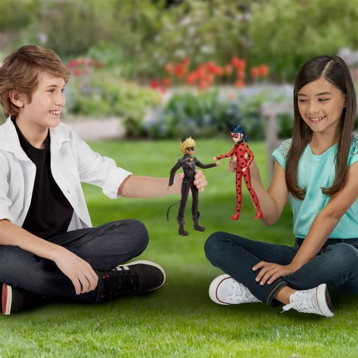 Zwei Kinder sitzen auf Gras und halten Puppen von Zeichentrickfiguren vor verschwommenem Hintergrund mit Grün und Blumen.