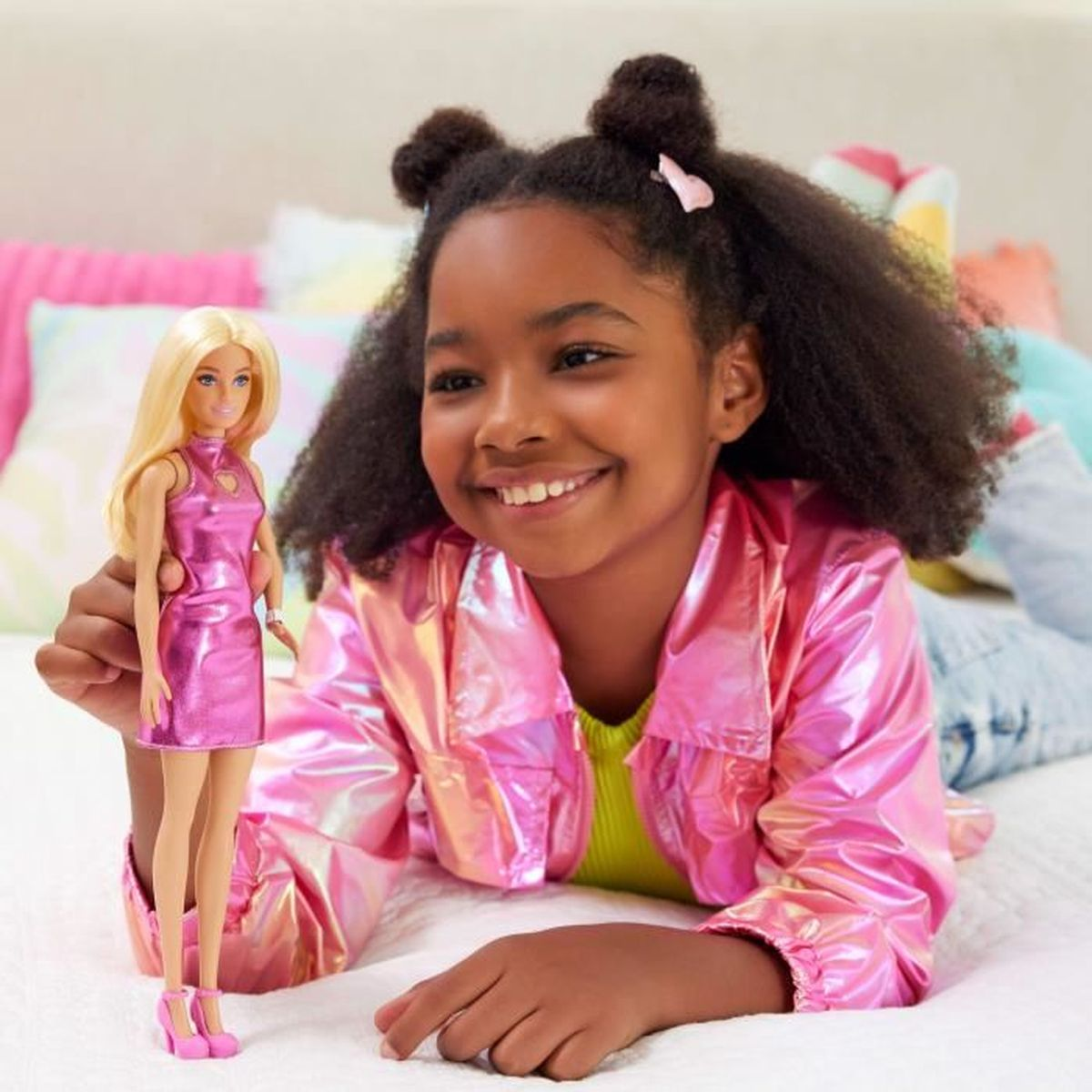 Ein lächelndes Mädchen hält eine Barbie-Puppe in einem rosa Kleid. Beide liegen auf einem Bett mit bunten Kissen. Das Mädchen trägt eine rosa Jacke.