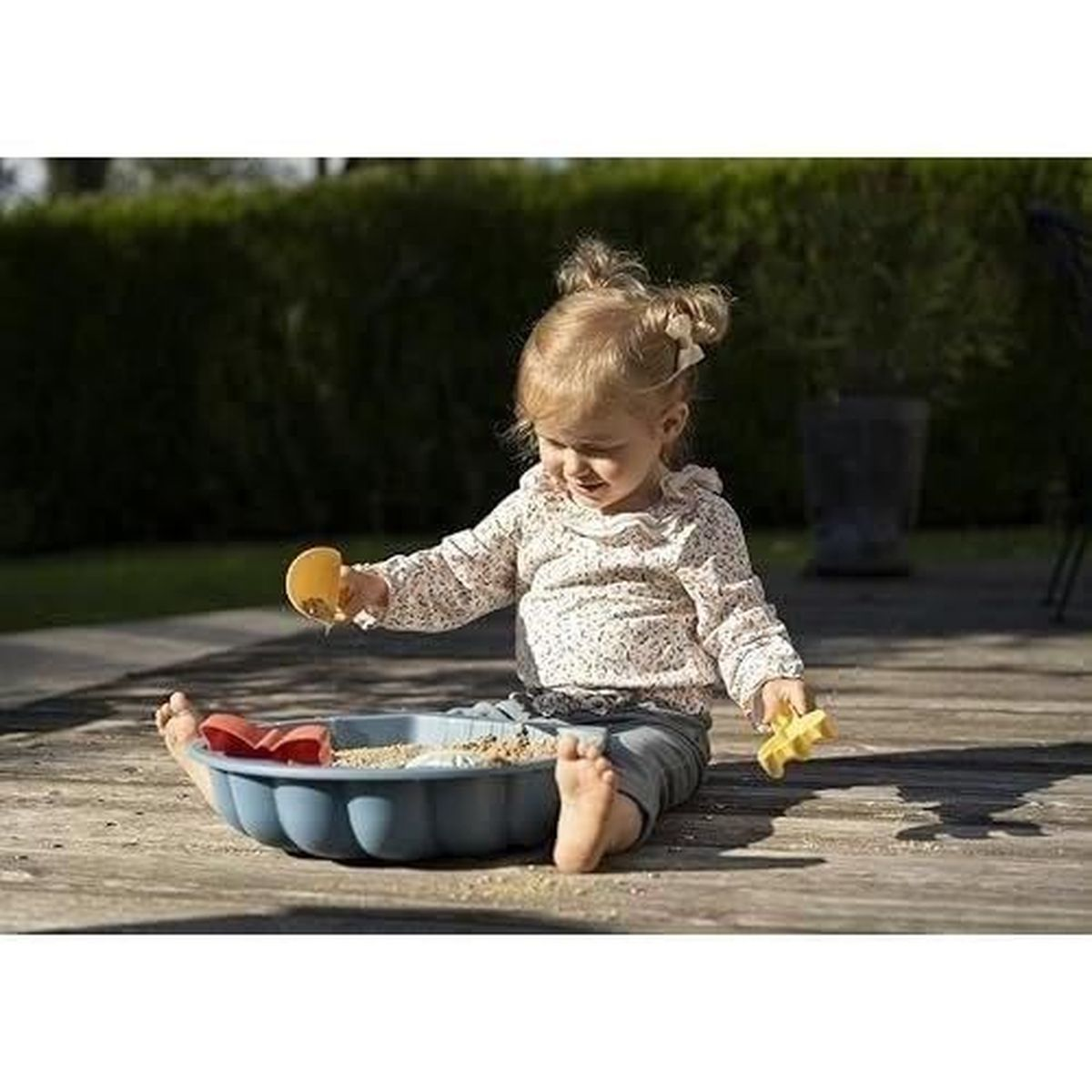 Un bambino gioca con sabbia e giocattoli in una sabbiera blu. Indossa una camicia floreale e pantaloncini, seduto su una terrazza di legno.