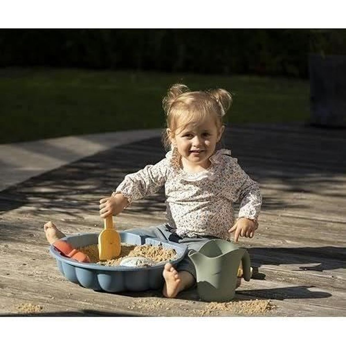 Una bambina gioca in una sabbiera con una paletta e un annaffiatoio. È seduta su un ponte di legno. C'è sabbia dappertutto.