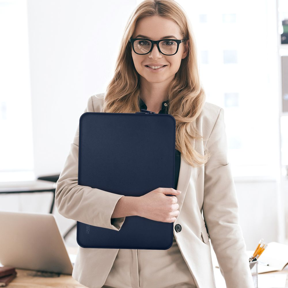 Blonde vrouw met bril houdt een marineblauwe laptoptas vast, lachend. Ze draagt een beige blazer. Een laptop en kantoorbenodigdheden zijn zichtbaar.