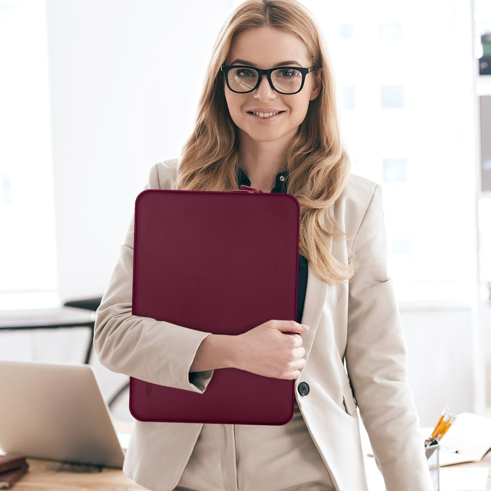 Een vrouw met een bril en blond haar houdt een bordeauxrode laptoptas vast. Ze draagt een beige blazer en glimlacht op kantoor.