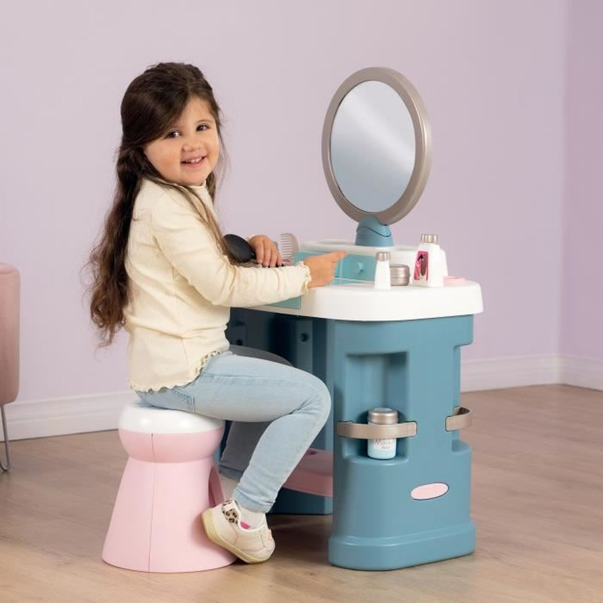 Ein Mädchen sitzt lächelnd an einem Spielzeugschminktisch. Der Schminktisch ist blau und weiß mit einem Spiegel. Vorne steht ein rosa Hocker.