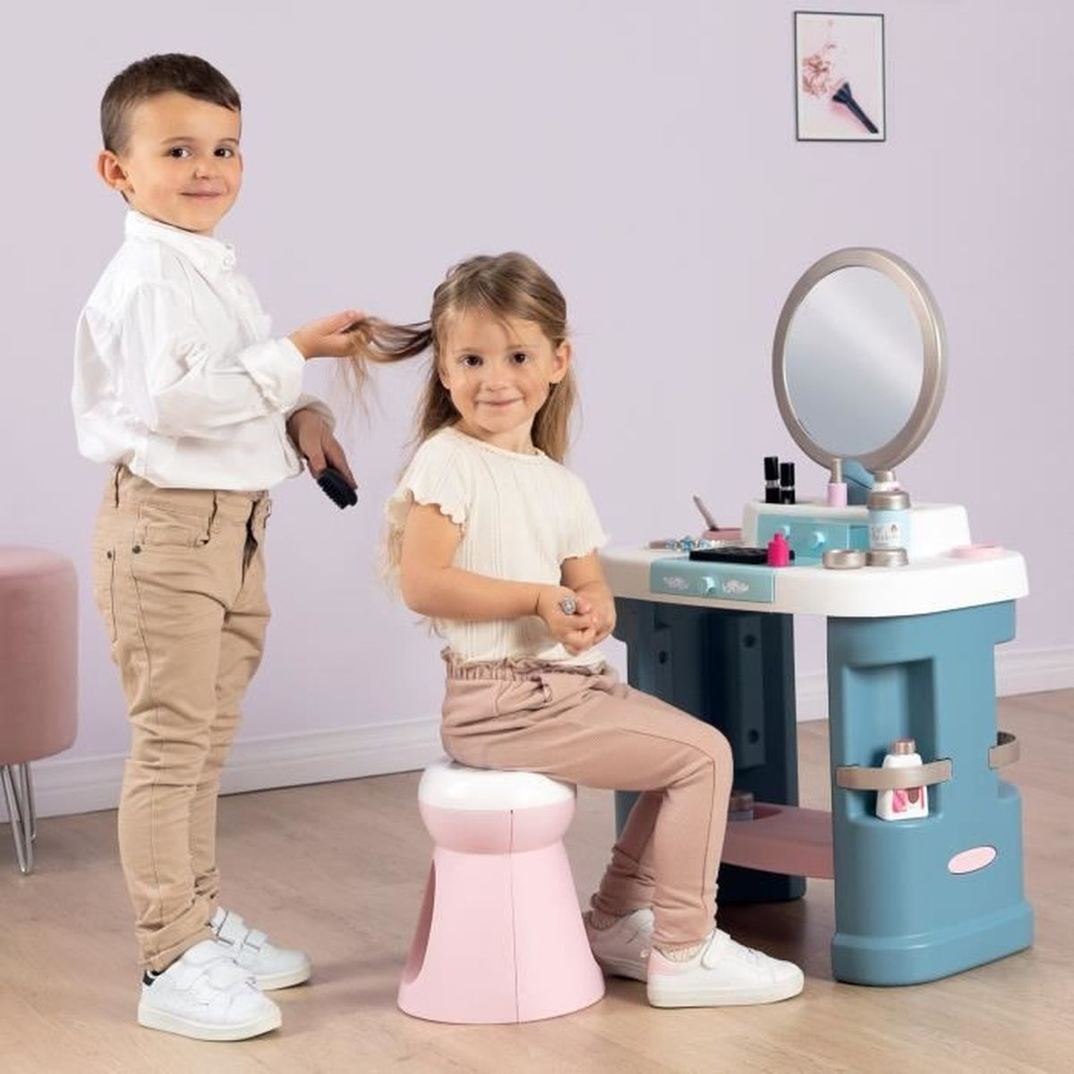 Ein Junge kämmt dem Mädchen die Haare. Sie stehen an einem Spielzeugschminktisch. Das Mädchen sitzt auf einem rosa Hocker.
