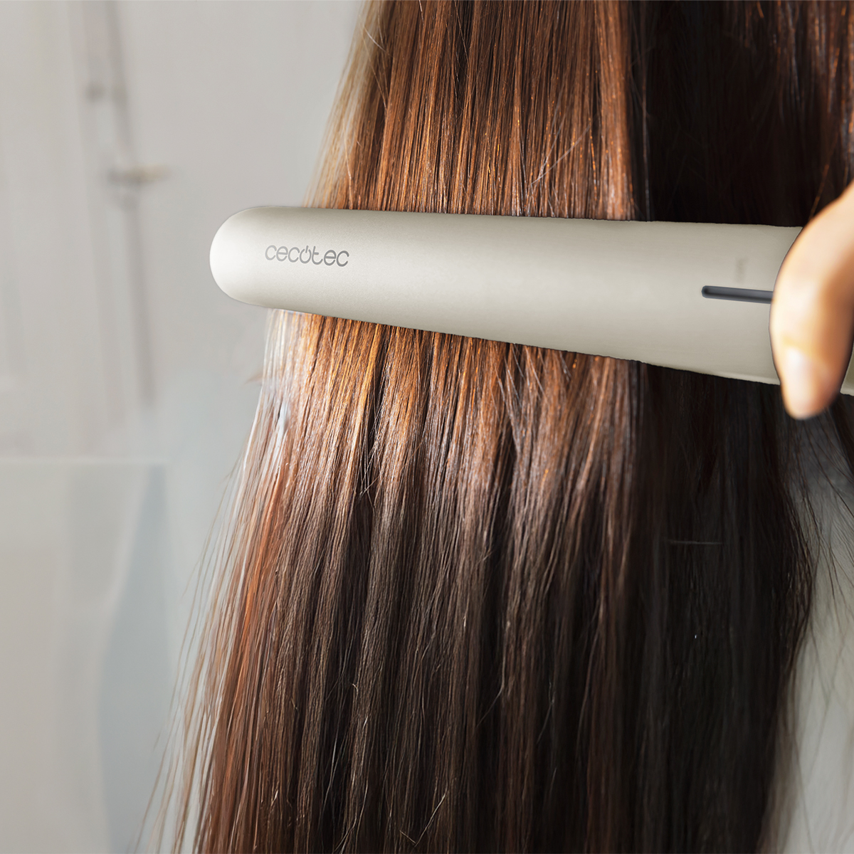Una persona usa una plancha de pelo en cabello largo y castaño. La plancha es beige y el fondo está borroso.