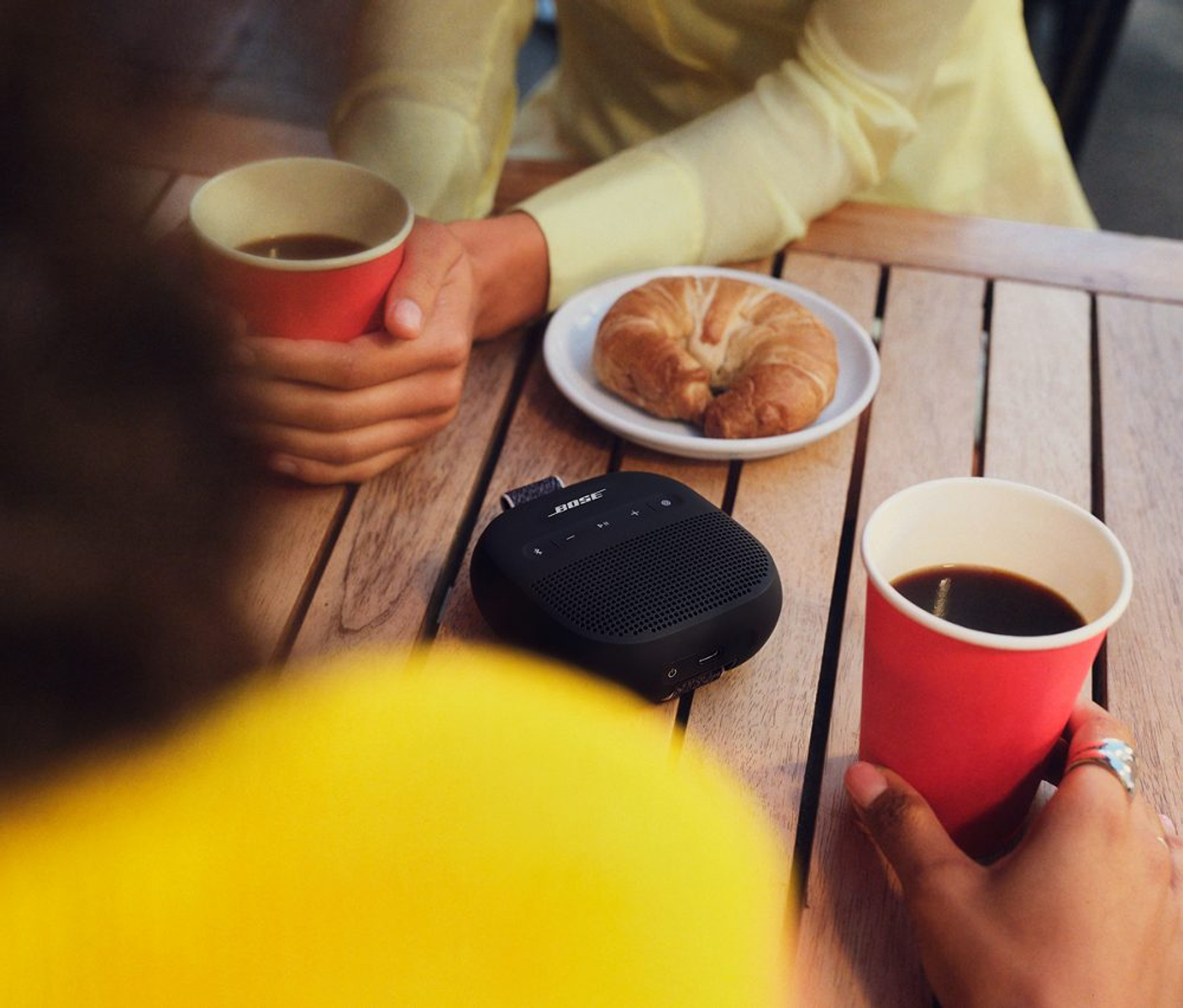 Dos personas en una mesa con café, croissant y un altavoz Bose. Una persona sostiene café. Se ven tops amarillos.