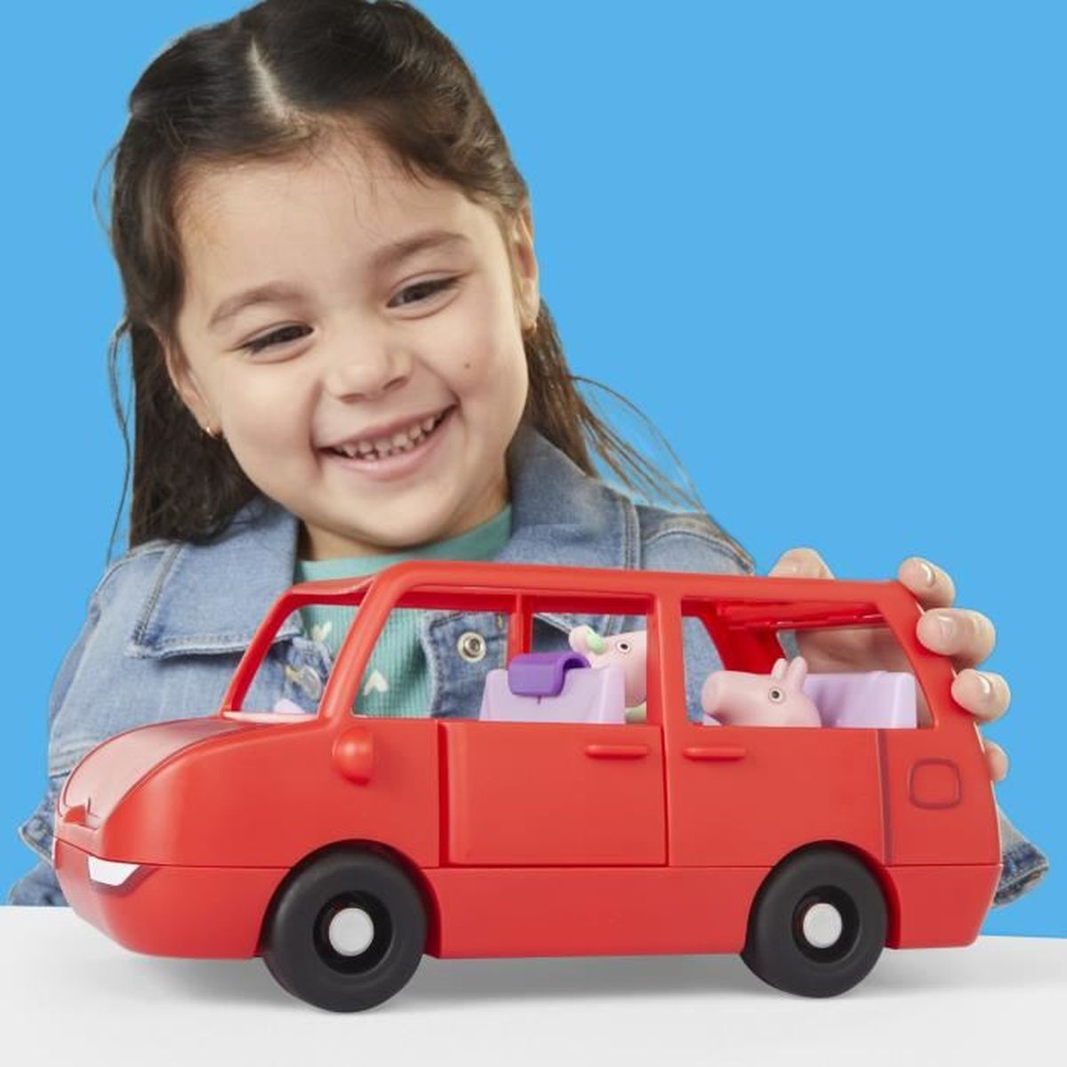 Una niña sonriente sostiene un coche de juguete rojo con dos figuras de cerdo dentro. El fondo es azul.