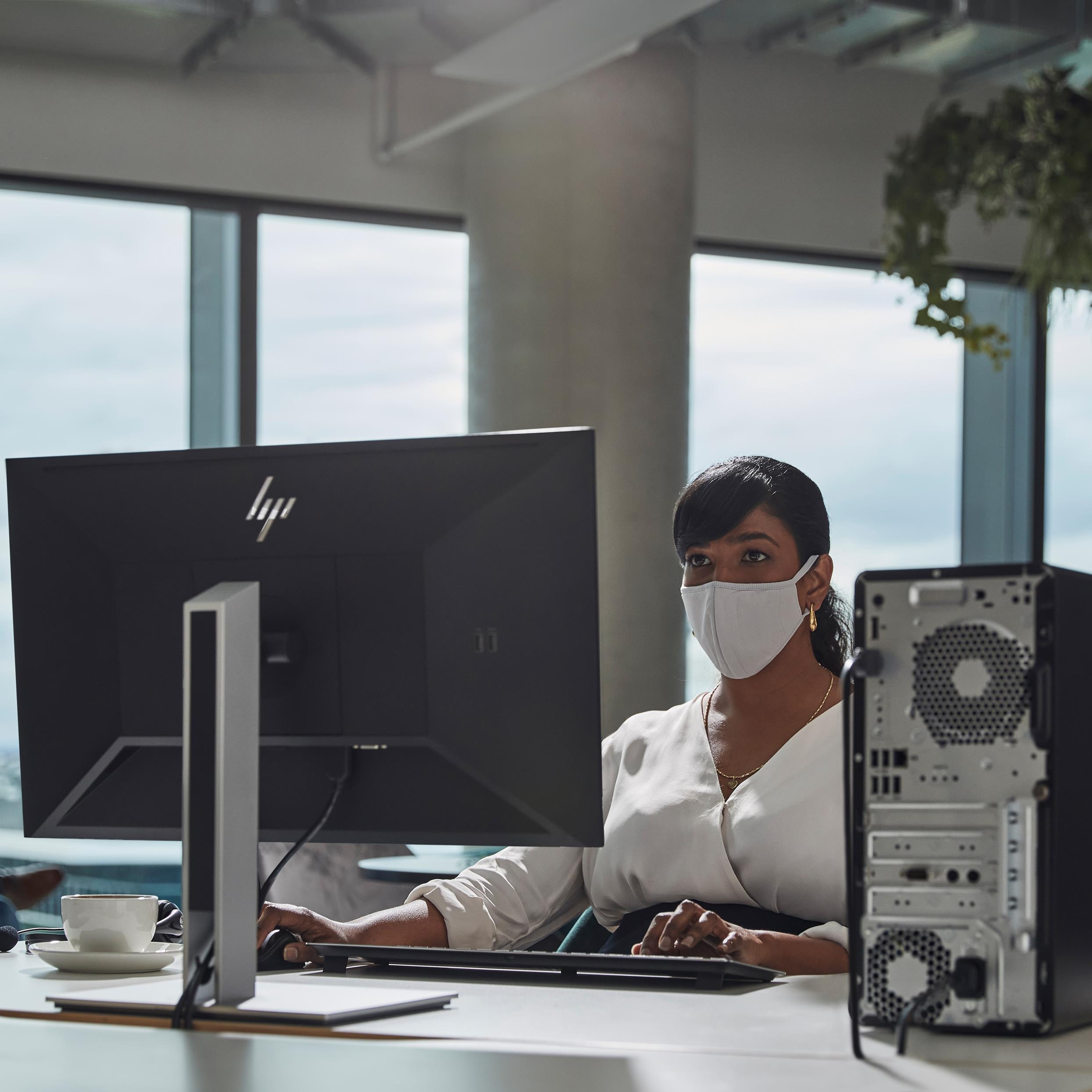 Una donna con la mascherina lavora al computer. Sul tavolo ci sono una tazza di caffè e un computer desktop.
