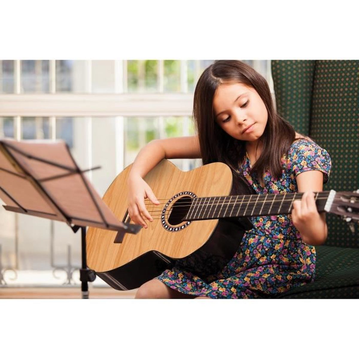Una niña toca una guitarra acústica de madera, mirando las cuerdas. Un atril está a la izquierda. Lleva un vestido floral.