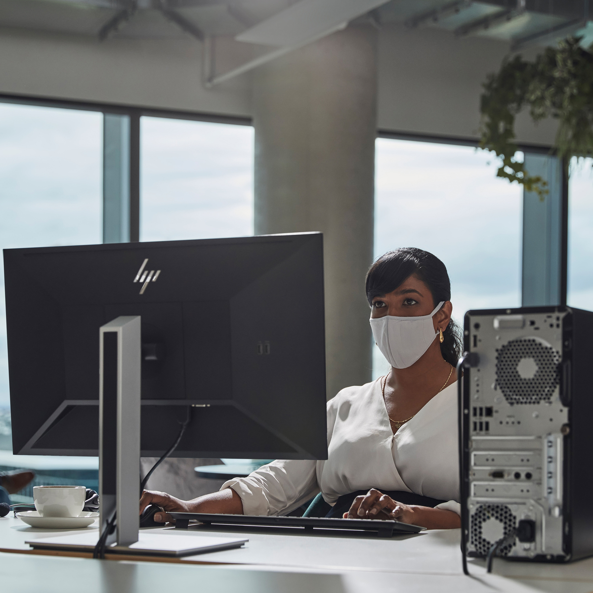 Una mujer con mascarilla trabaja en una computadora. Se ven un monitor negro y una torre de computadora. Está en una oficina.