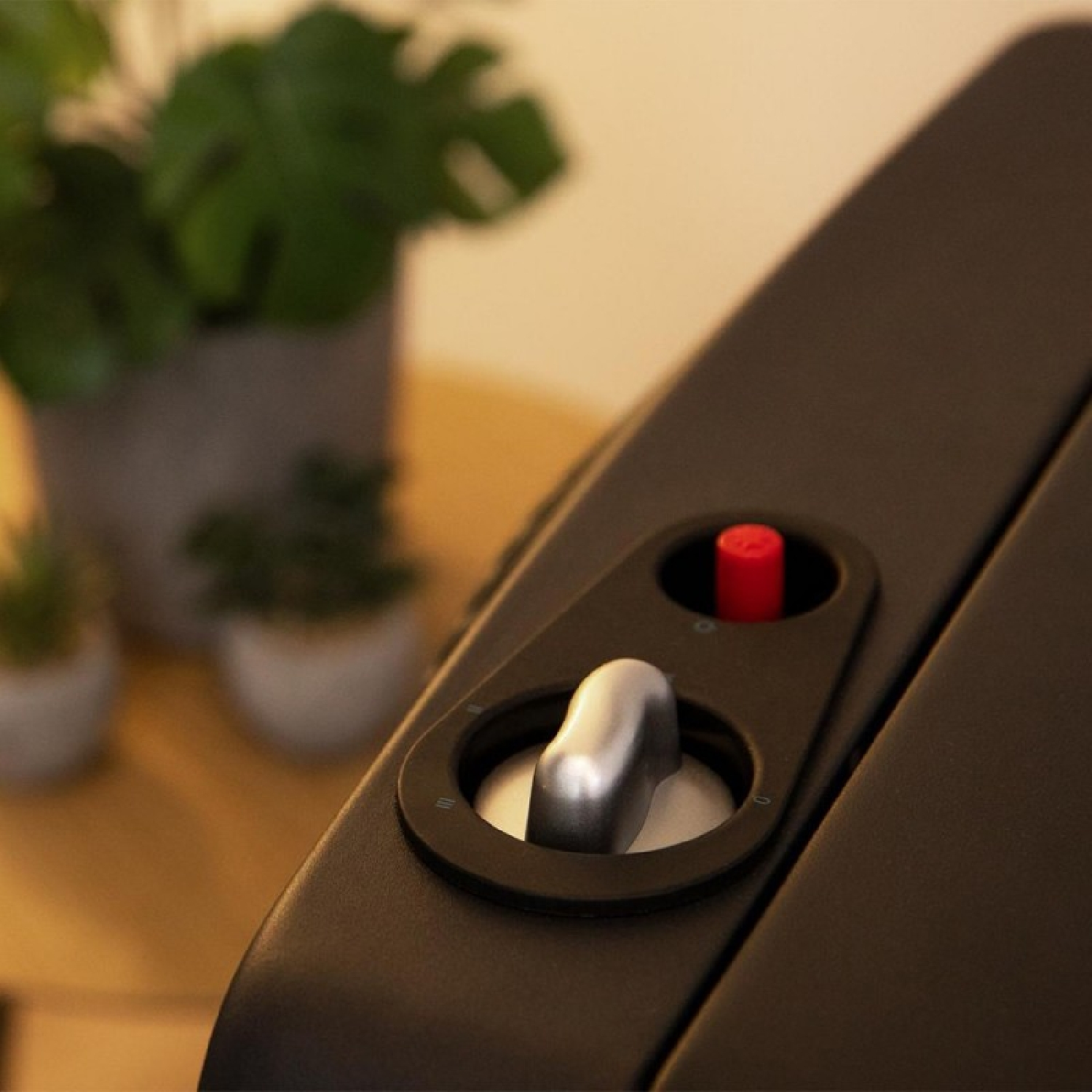 Primo piano di un dispositivo nero con una manopola argentata e un pulsante rosso. Piante verdi sullo sfondo, su un tavolo di legno.