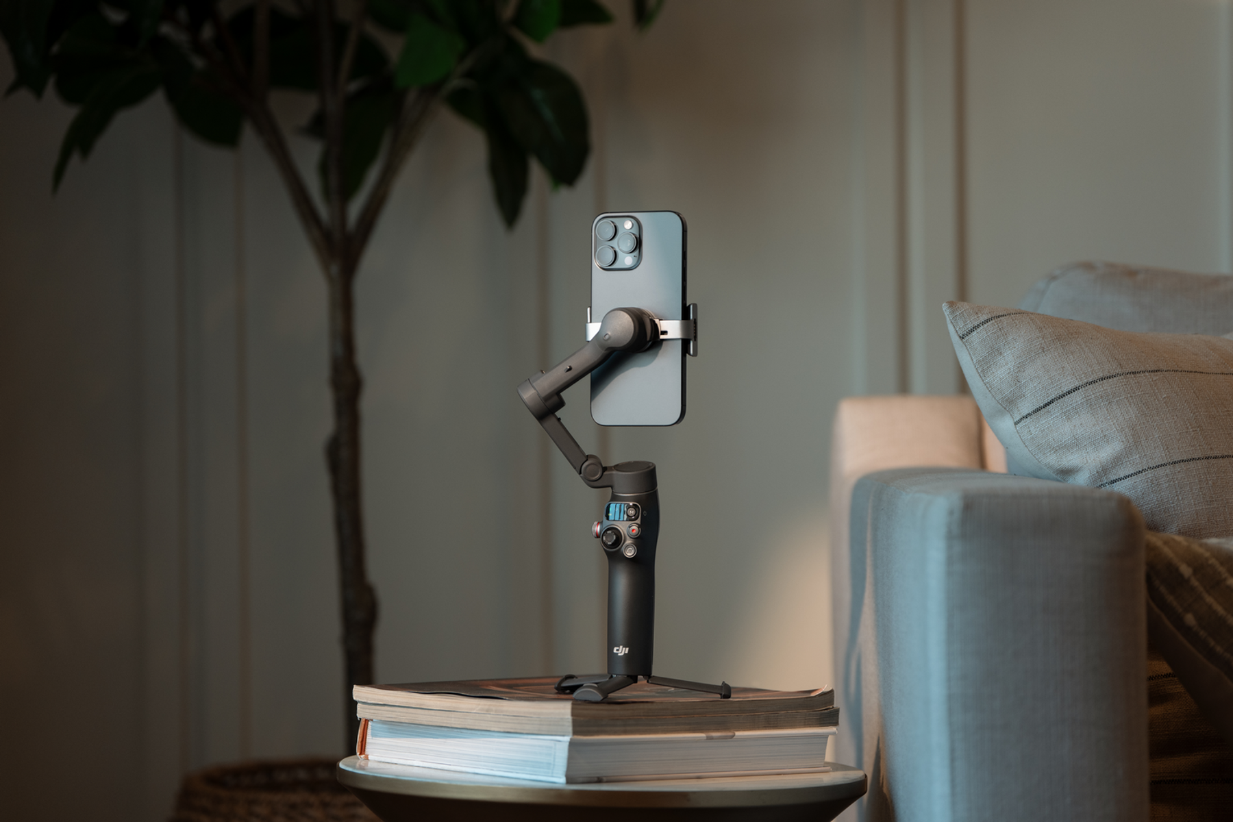 Smartfon jest zamontowany na czarnym gimbalu, umieszczonym na książkach. Roślina i sofa są w tle.