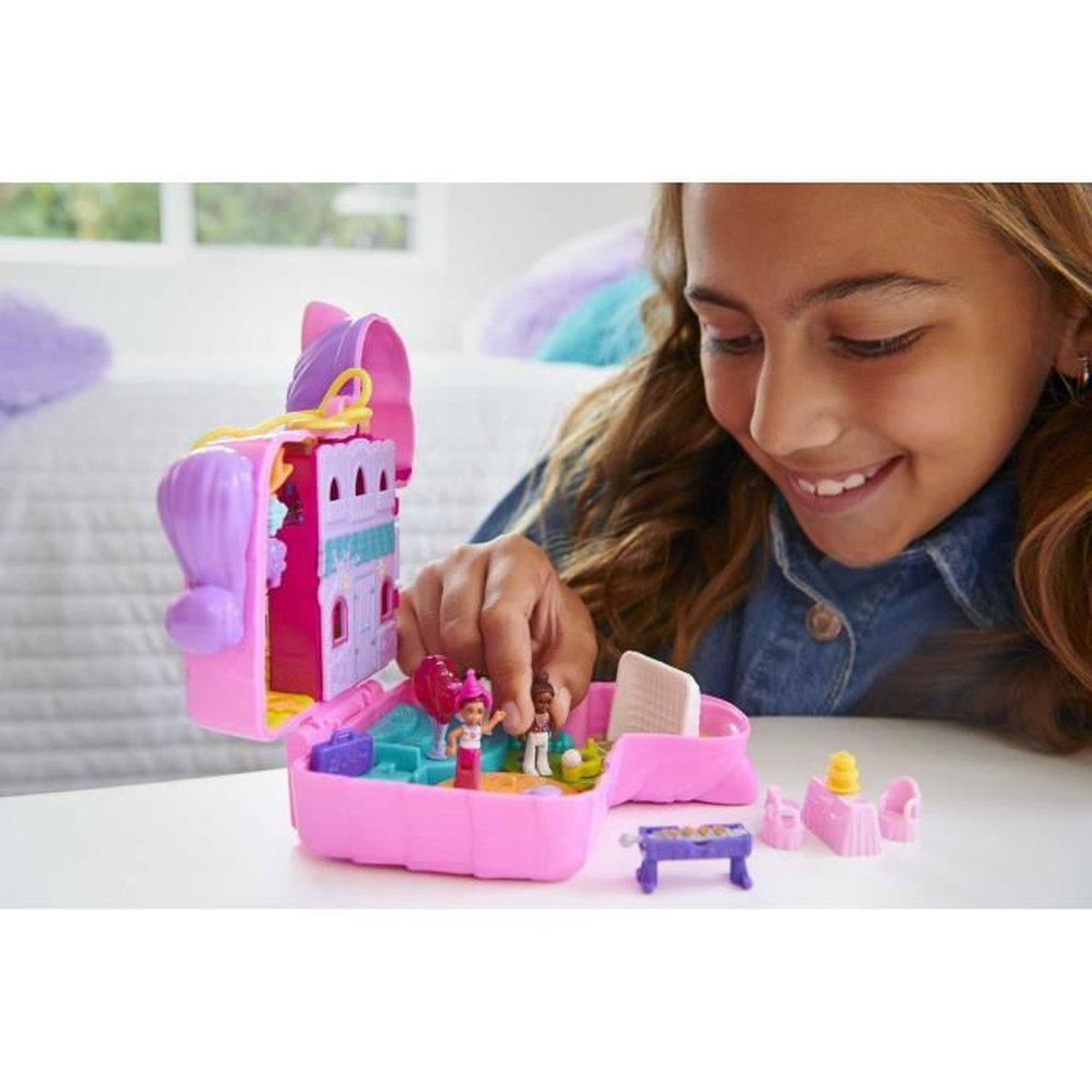Ein Mädchen spielt mit einem rosa Polly Pocket-Spielzeug. Das Spielzeug ist geöffnet und zeigt Miniaturfiguren, Möbel und ein Schloss. Das Mädchen lächelt.