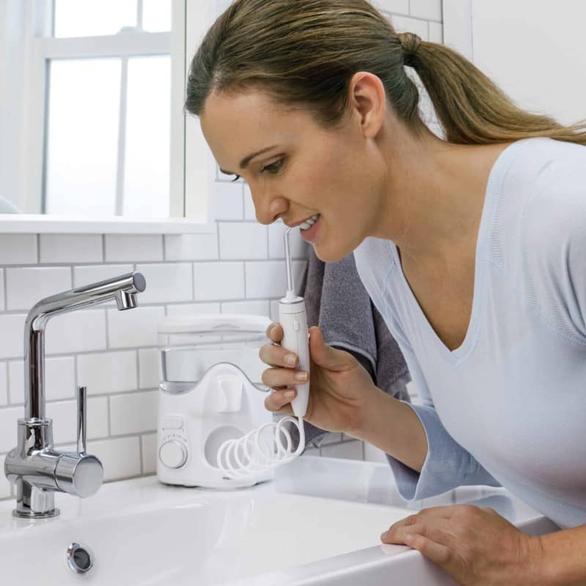 Una donna usa un idropulsore dentale in bagno. Tiene il dispositivo vicino alla bocca, accanto a un lavandino, con un'unità.
