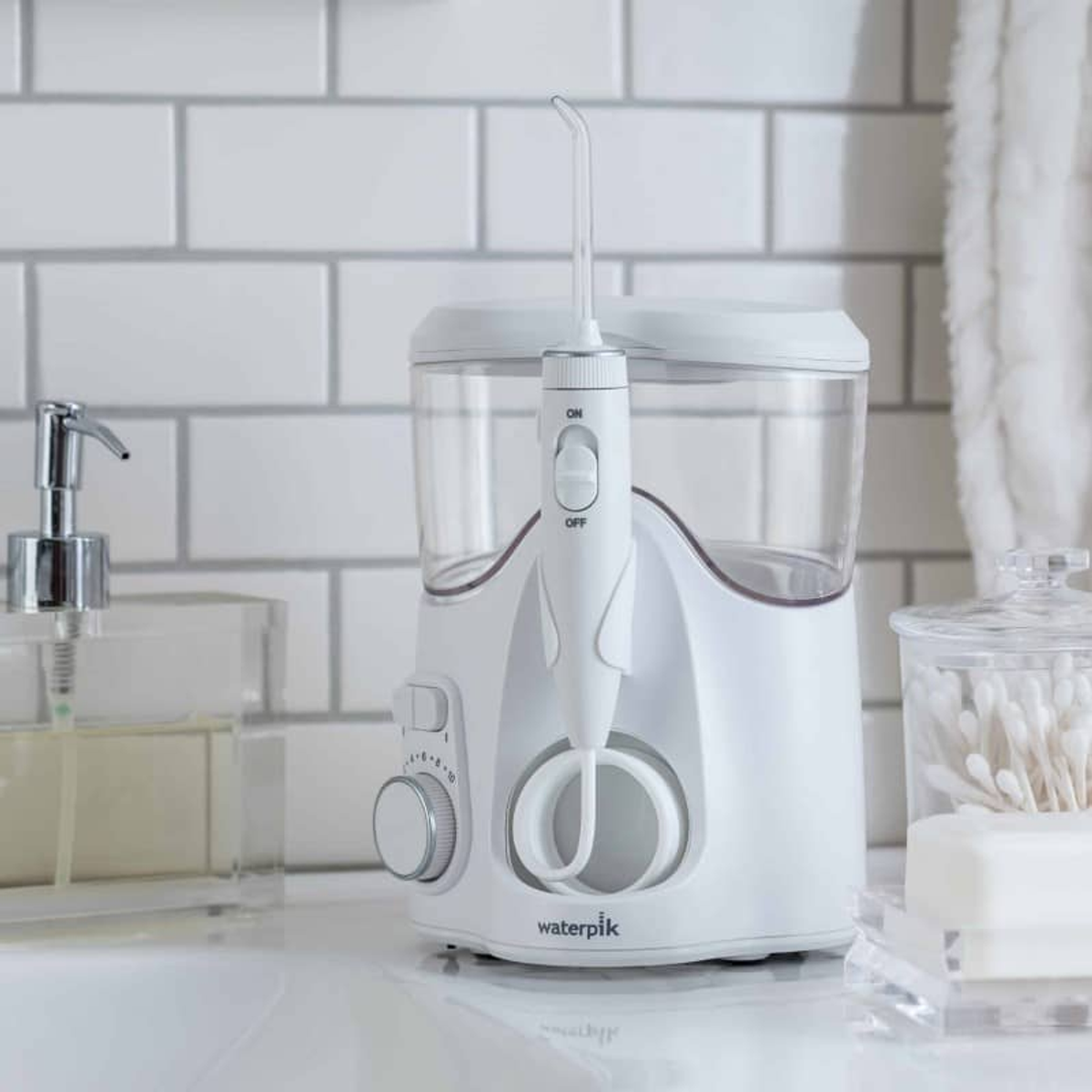Un idropulsore Waterpik bianco è appoggiato su un bancone. Sapone, cotton fioc e un dosatore di sapone sono visibili anche in bagno.