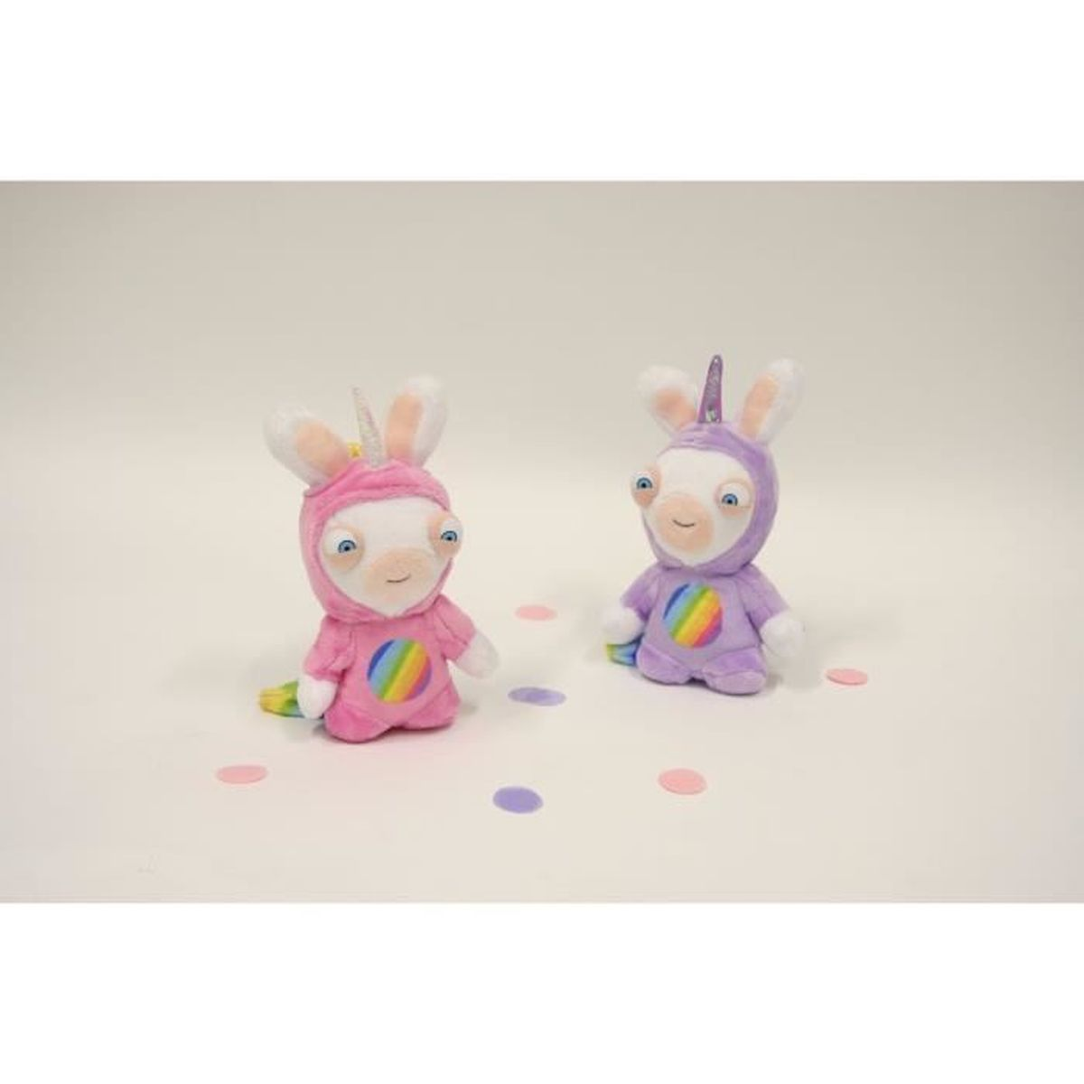Zwei Stoffhasen, einer rosa und einer lila, tragen Einhörner-Kostüme mit Regenbogenakzenten auf einer weißen Oberfläche mit Konfetti.