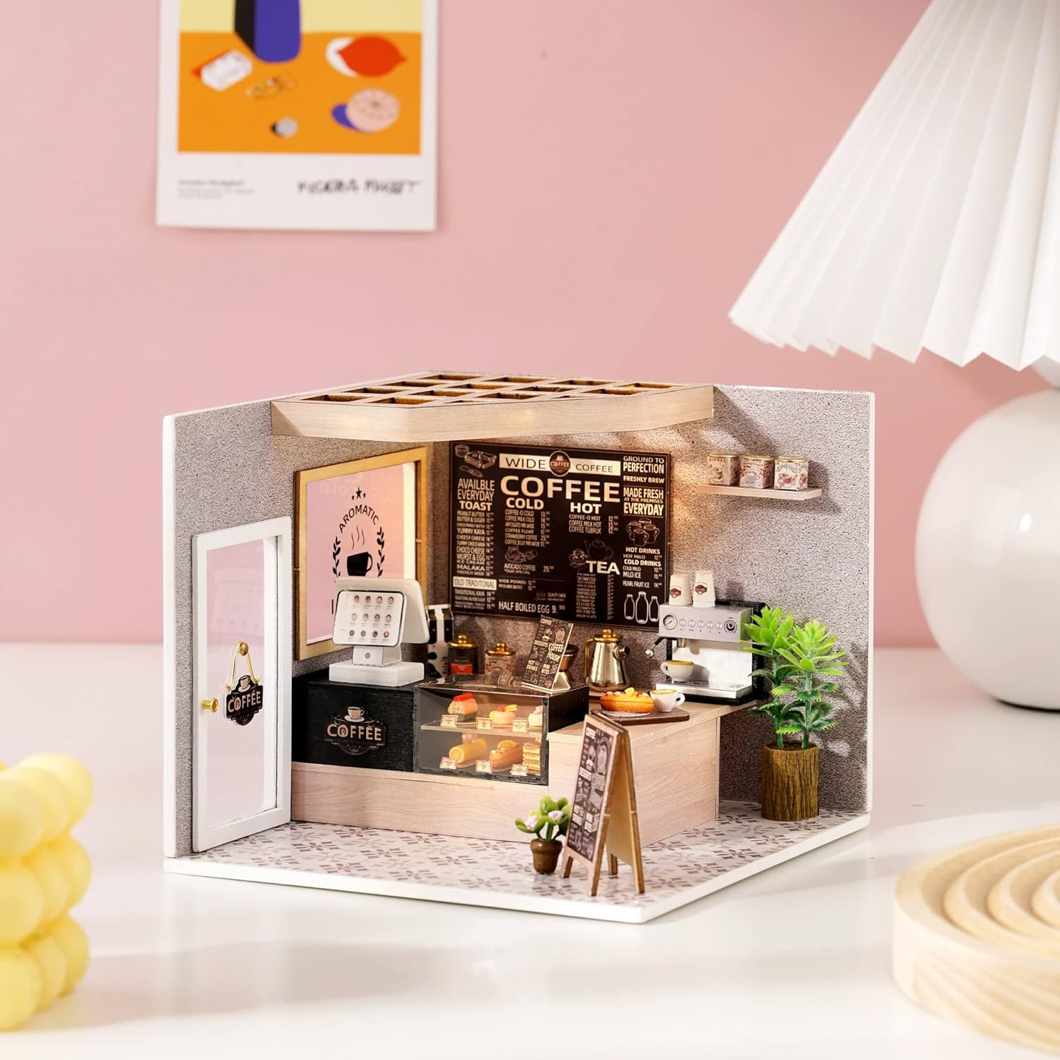 Miniatur-Kaffeehausmodell. Es hat eine Kaffeekarte, Gebäck und eine Kaffeemaschine. Eine kleine Pflanze und eine Lampe sind ebenfalls vorhanden.