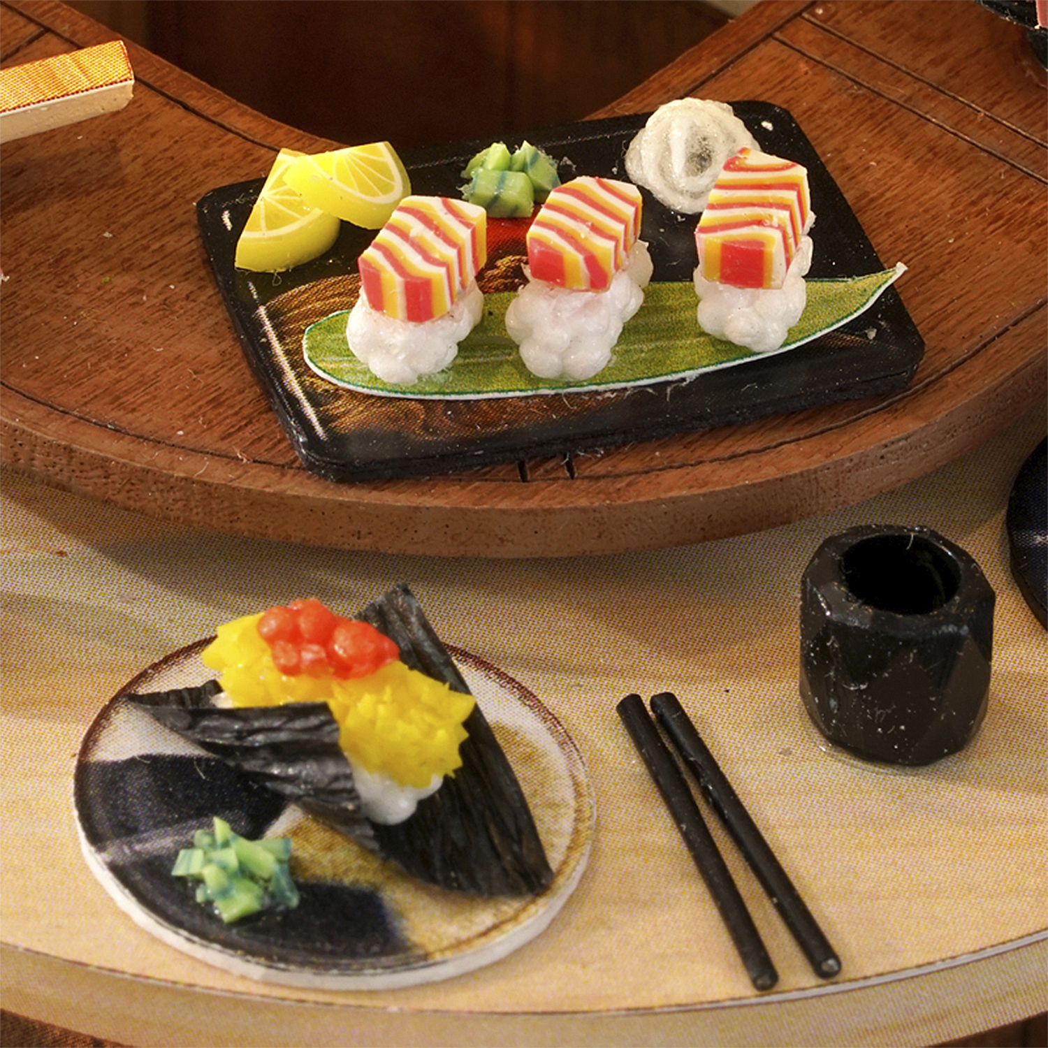 Set di sushi in miniatura. Include sushi su un vassoio, un piattino con sushi, bacchette e un contenitore nero.