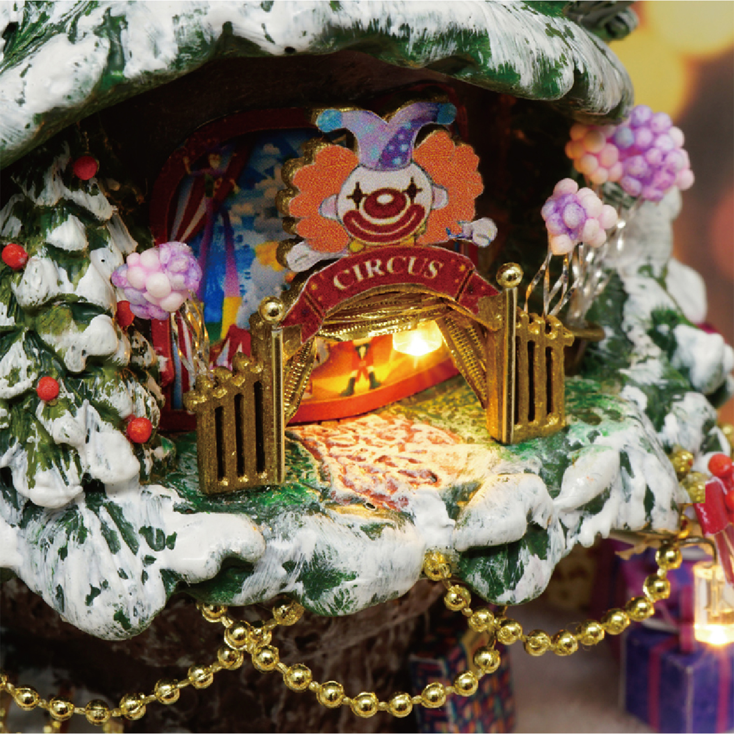 Una scena circense in miniatura con un clown, un'insegna del circo, alberi innevati e una ghirlanda di perline dorate. Il circo è illuminato.