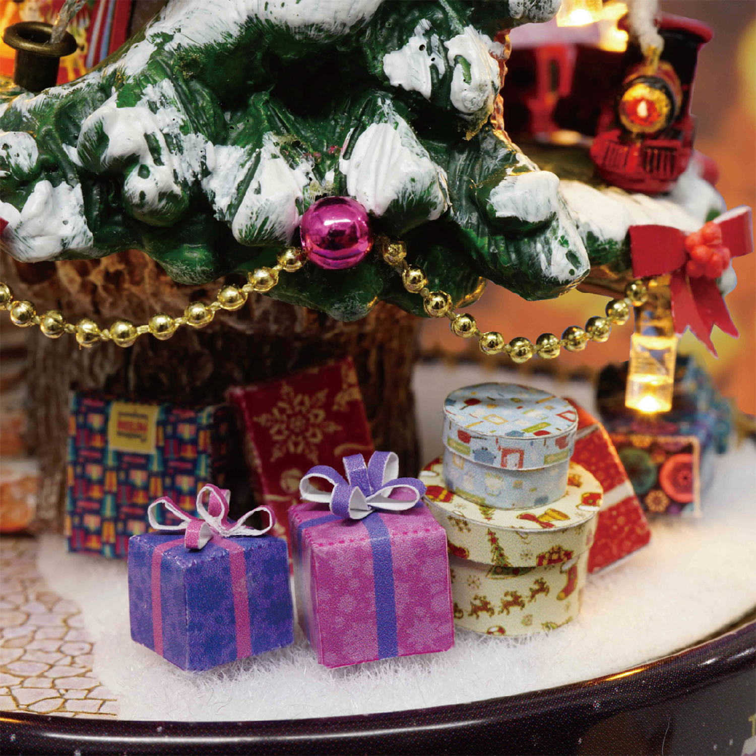 Una scena natalizia in miniatura con regali, un albero e un piccolo treno. L'albero è coperto di neve, con un ornamento rosa e perline dorate.