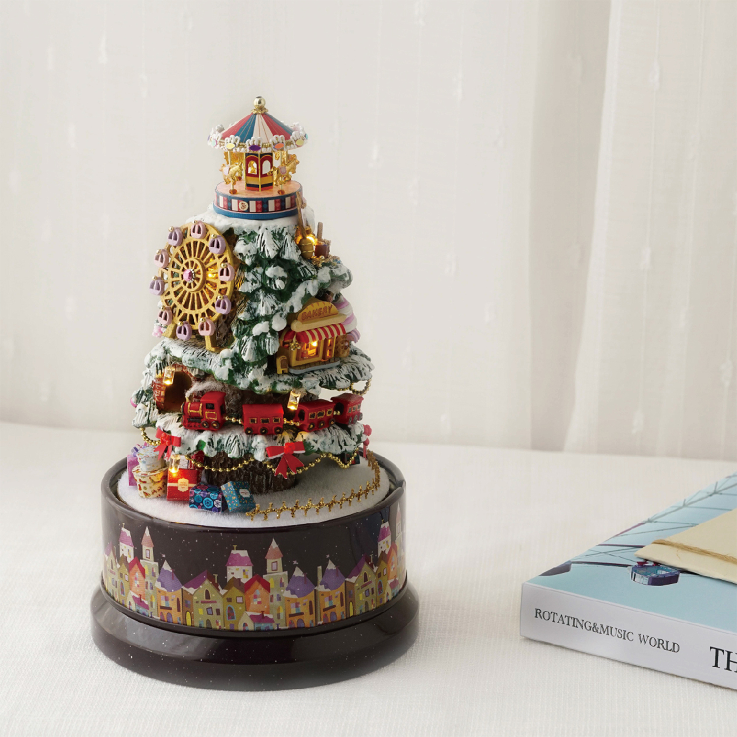 Un ornamento per albero di Natale con giostra, ruota panoramica e treno. Un libro è a destra. Tende bianche sullo sfondo.