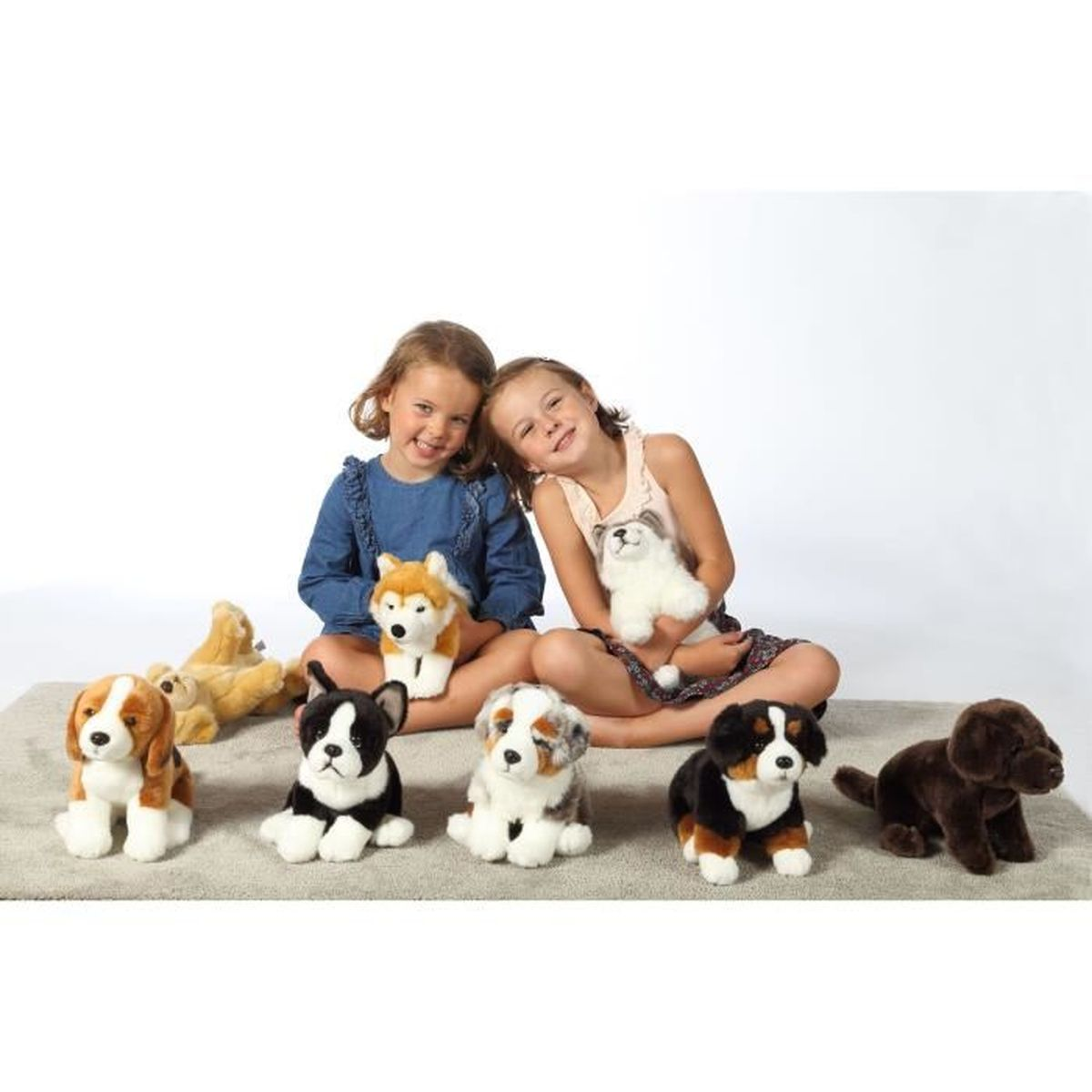 Dos niñas sonrientes sostienen perros de peluche. Varios perros de peluche están sentados en una alfombra frente a ellas, incluyendo un beagle y un perro marrón.