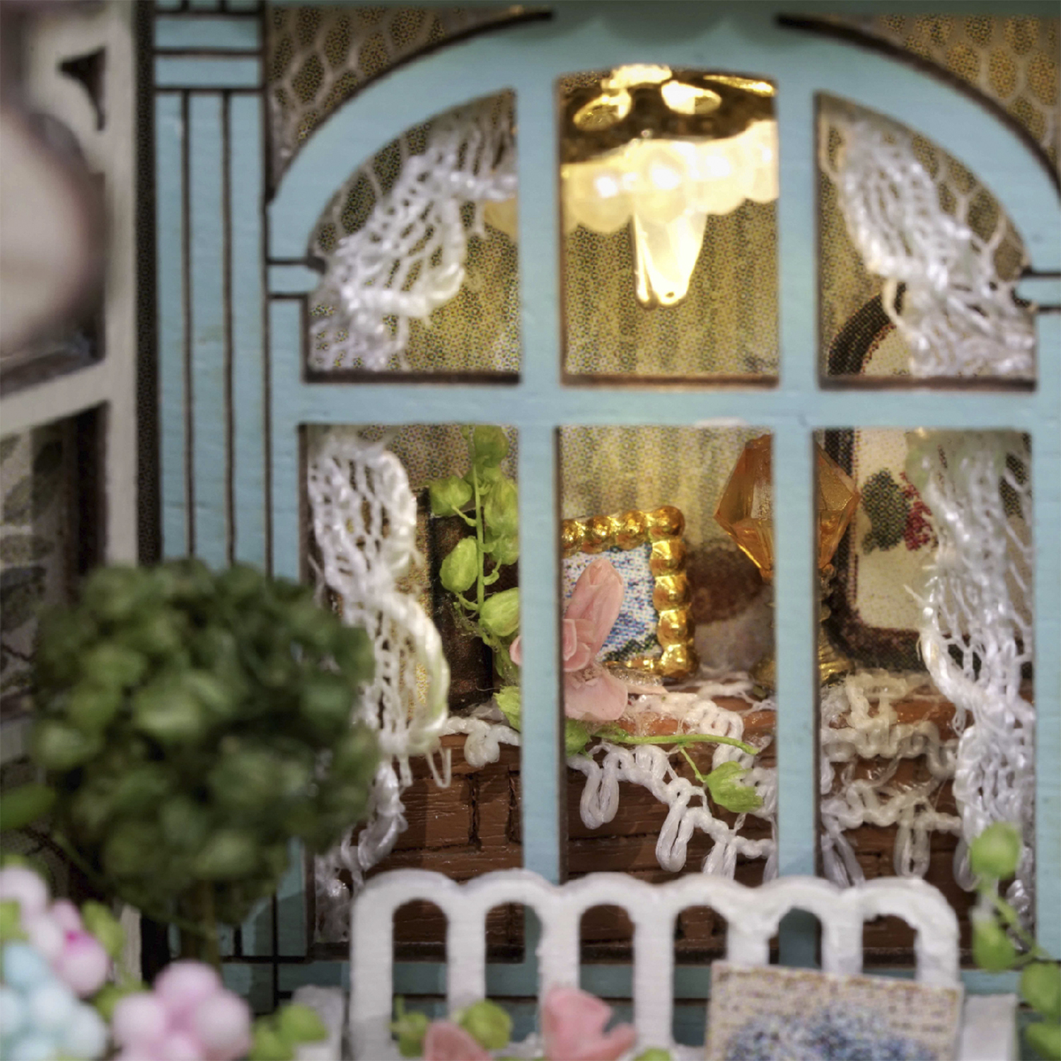 Scena di casa in miniatura. Una finestra blu incornicia una lampada illuminata. Tendine di pizzo, una piccola cornice e piante verdi sono visibili.