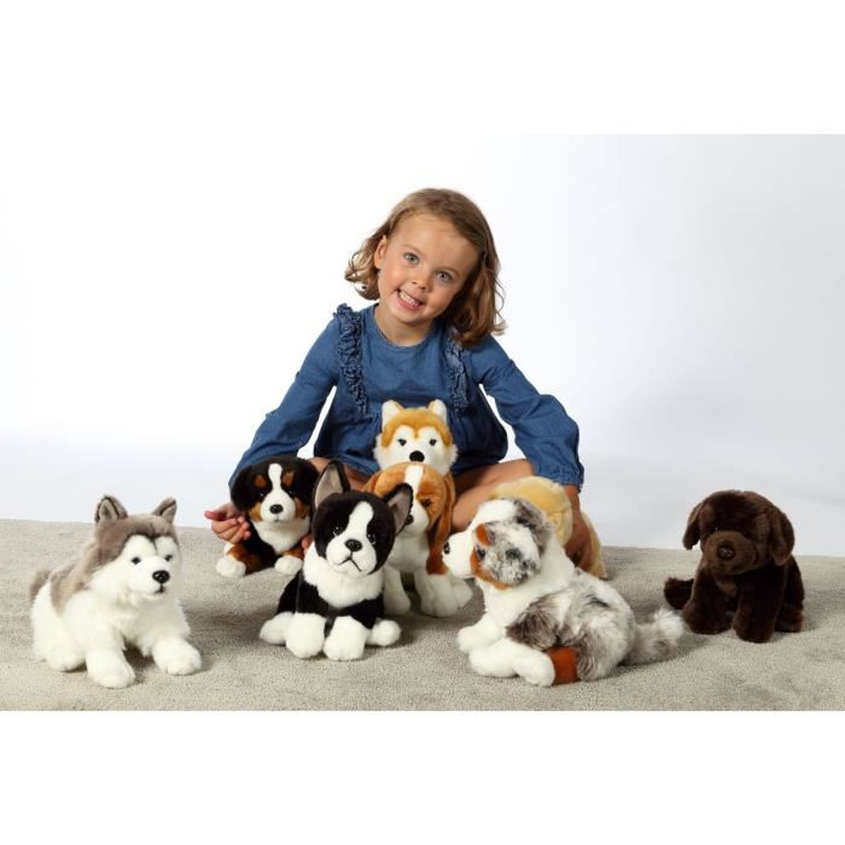 Una niña sonríe rodeada de peluches de perros. Hay varias razas presentes, incluyendo huskys y un labrador marrón. La escena es sobre una alfombra.