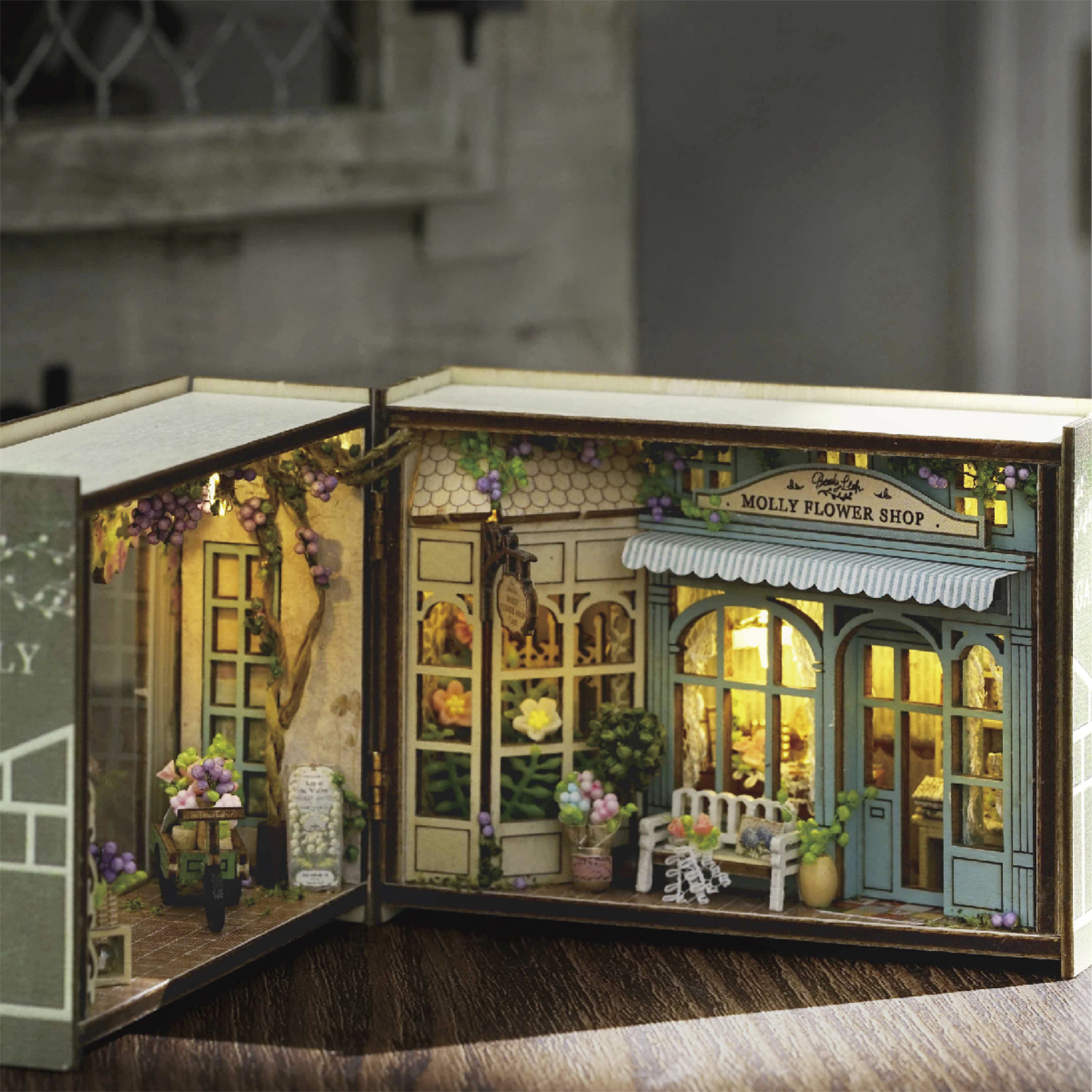Modello di negozio di fiori in miniatura. Ha una tenda da sole blu, una panchina e un cartello con scritto 'Molly Flower Shop'. Il modello è dentro un libro.