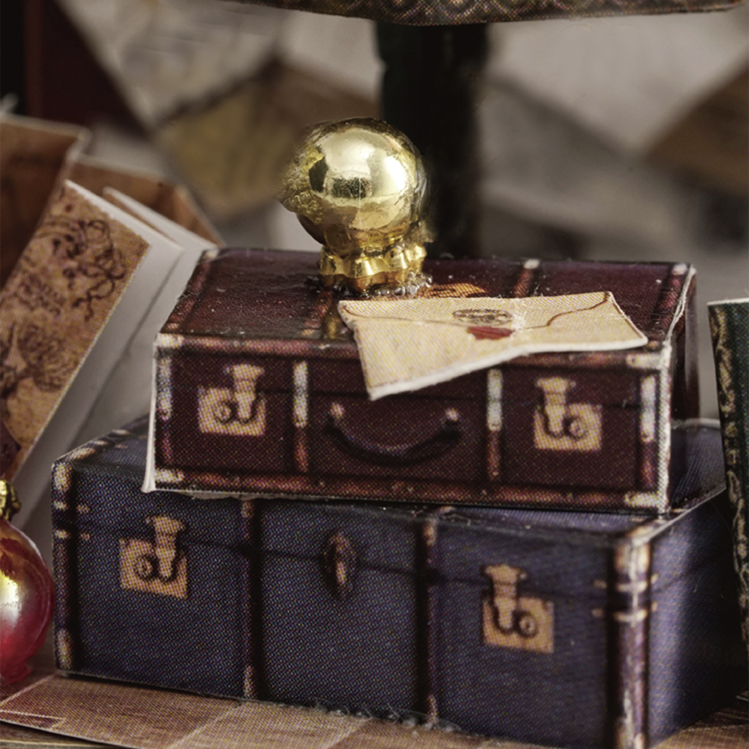 Valigie in miniatura impilate, sormontate da un globo dorato. Una lettera è appoggiata sulla valigia superiore. Dominano i toni blu scuro e marrone.